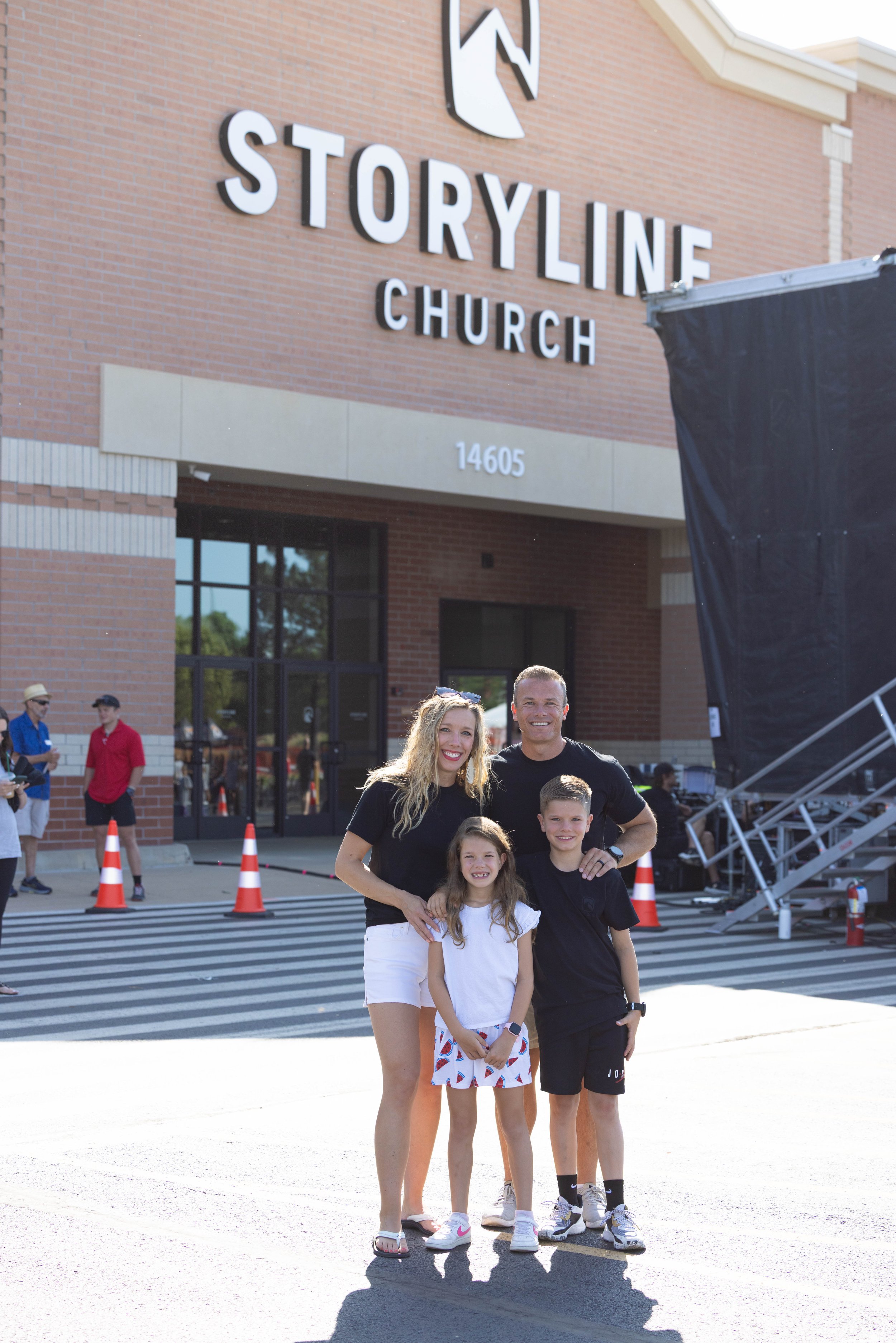 A family of four standing outside the Stone Church, smiling for a photo in front of the building's brick facade and large sign.