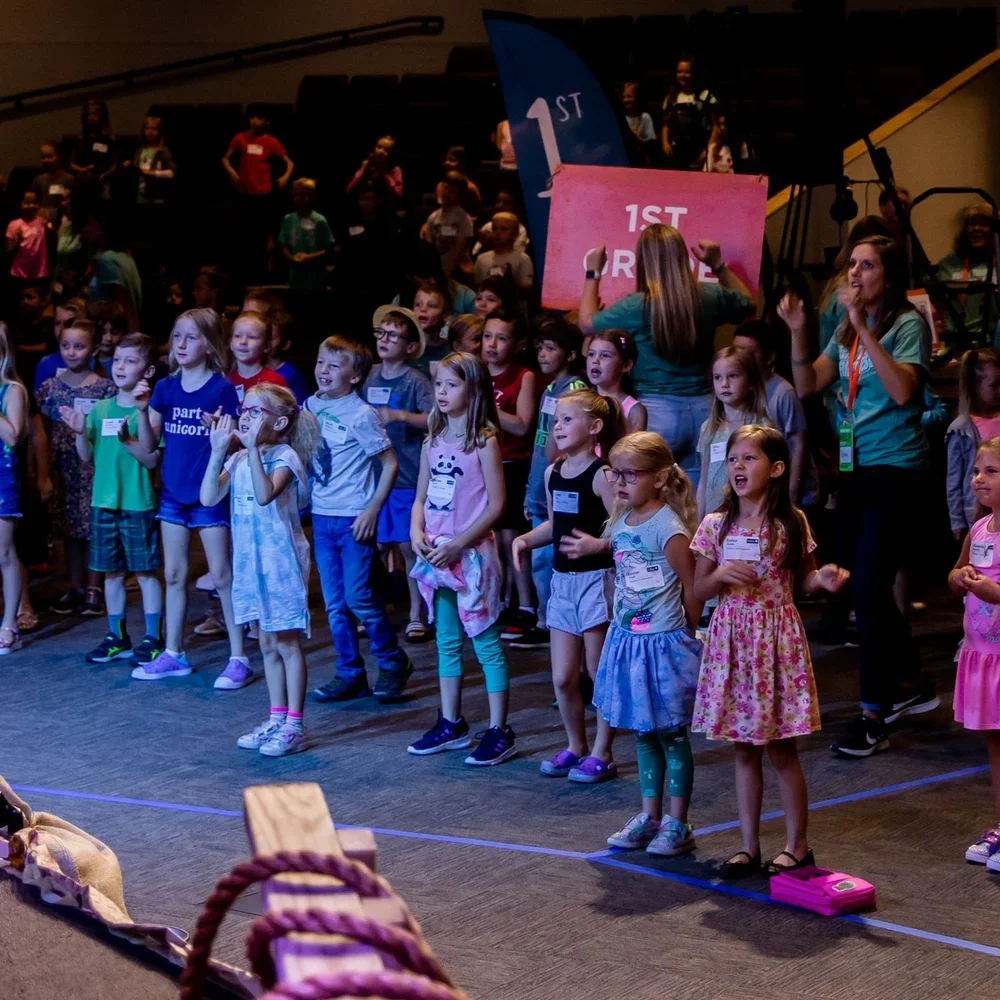 Children participating in a group activity on a stage, with adults assisting, some holding signs that say "1st" and "1st R"; children appear excited and engaged.