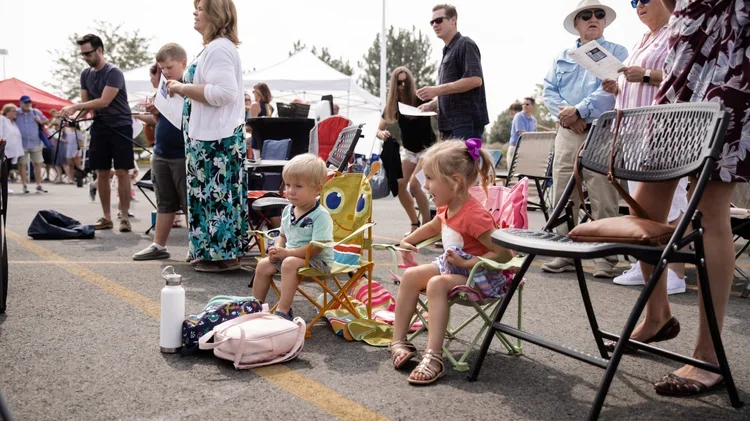 Children sitting in chairs at an outdoor event, surrounded by adults and tents, with some holding papers, during daytime.
