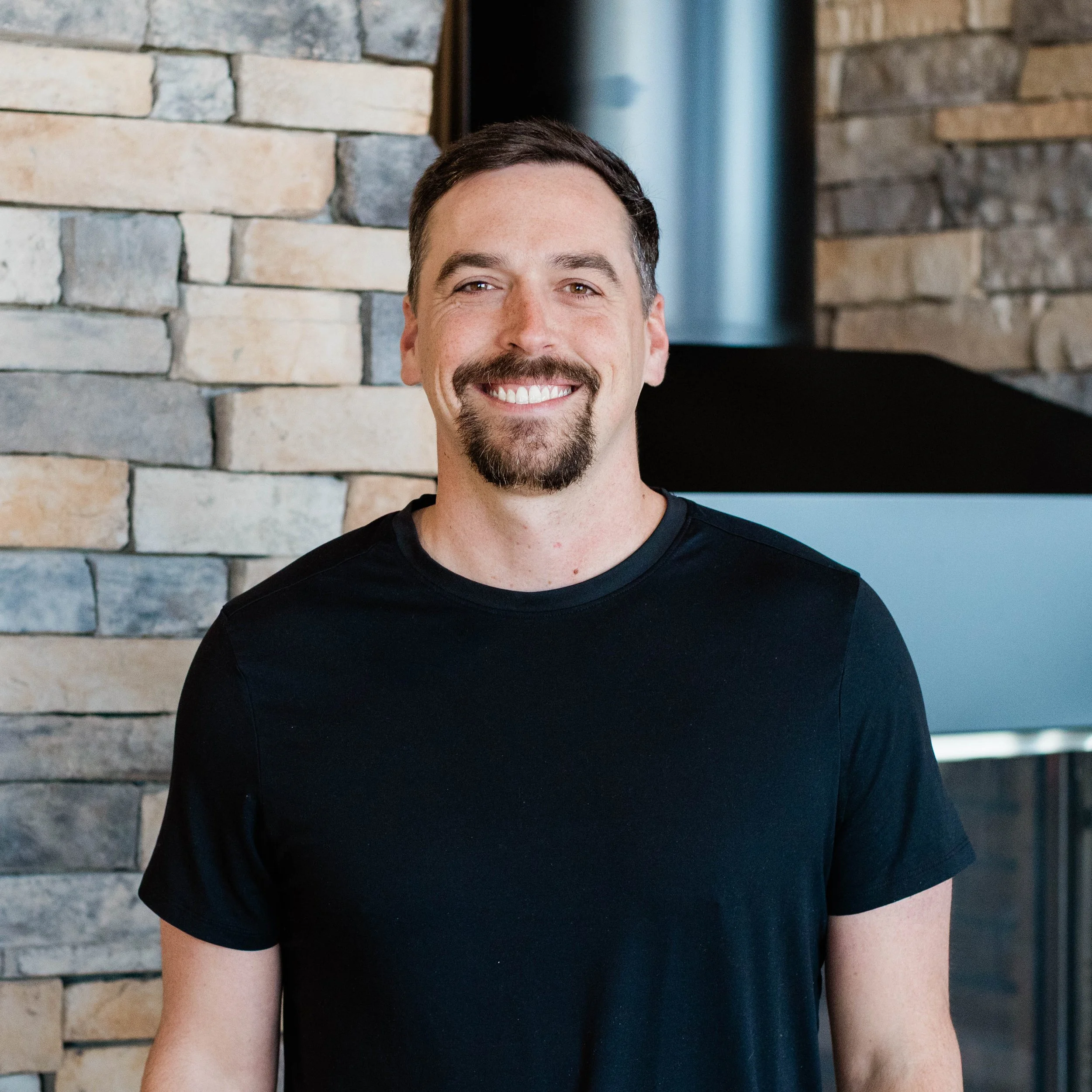A man with dark hair and a goatee smiling, wearing a navy Under Armour hoodie, standing in front of a textured stone wall.