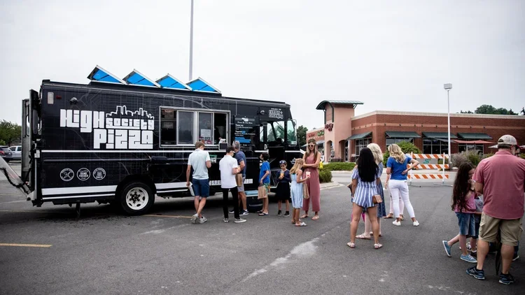 People lining up at a pizza food truck in a parking lot outside a building with a pink exterior