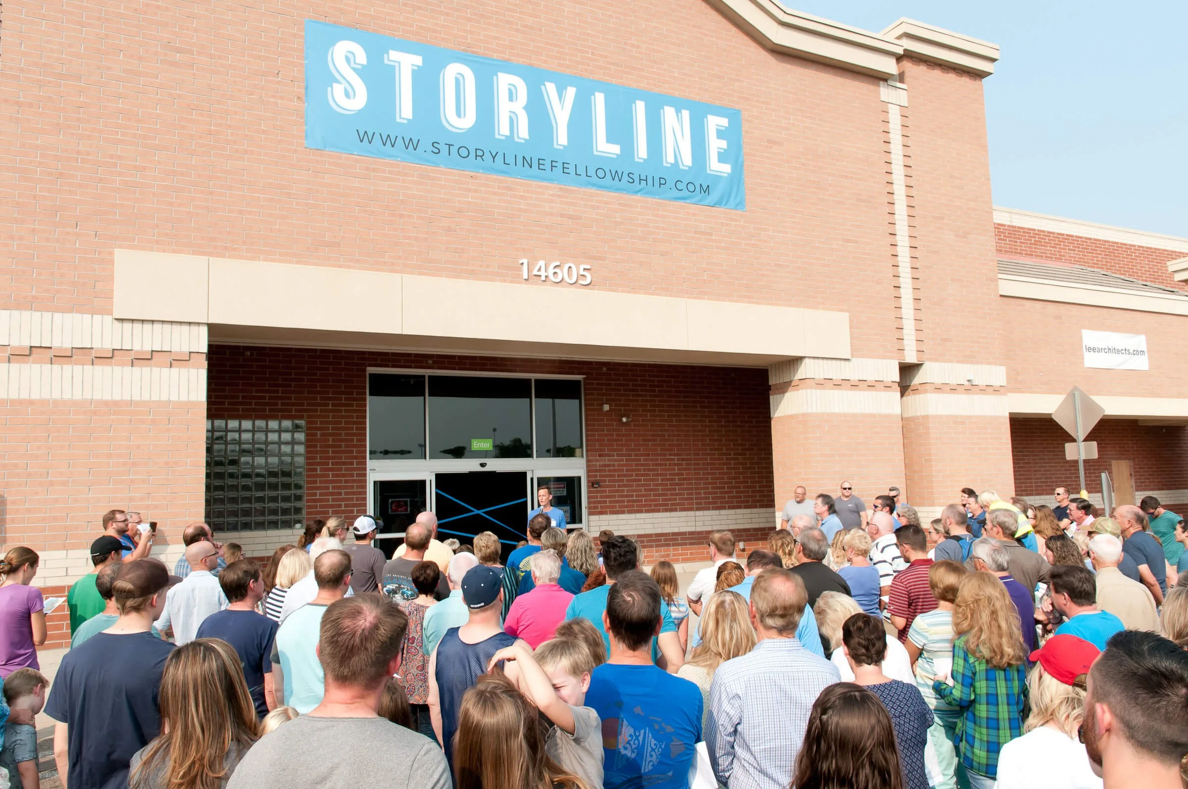 A crowd gathered outside a brick building for an event, with a large blue sign reading 'STORYLINE' and the text 'SUMMER 2018' overlaid. People are listening to a speaker at a podium, some taking photos, with the entrance door visible behind the speaker.