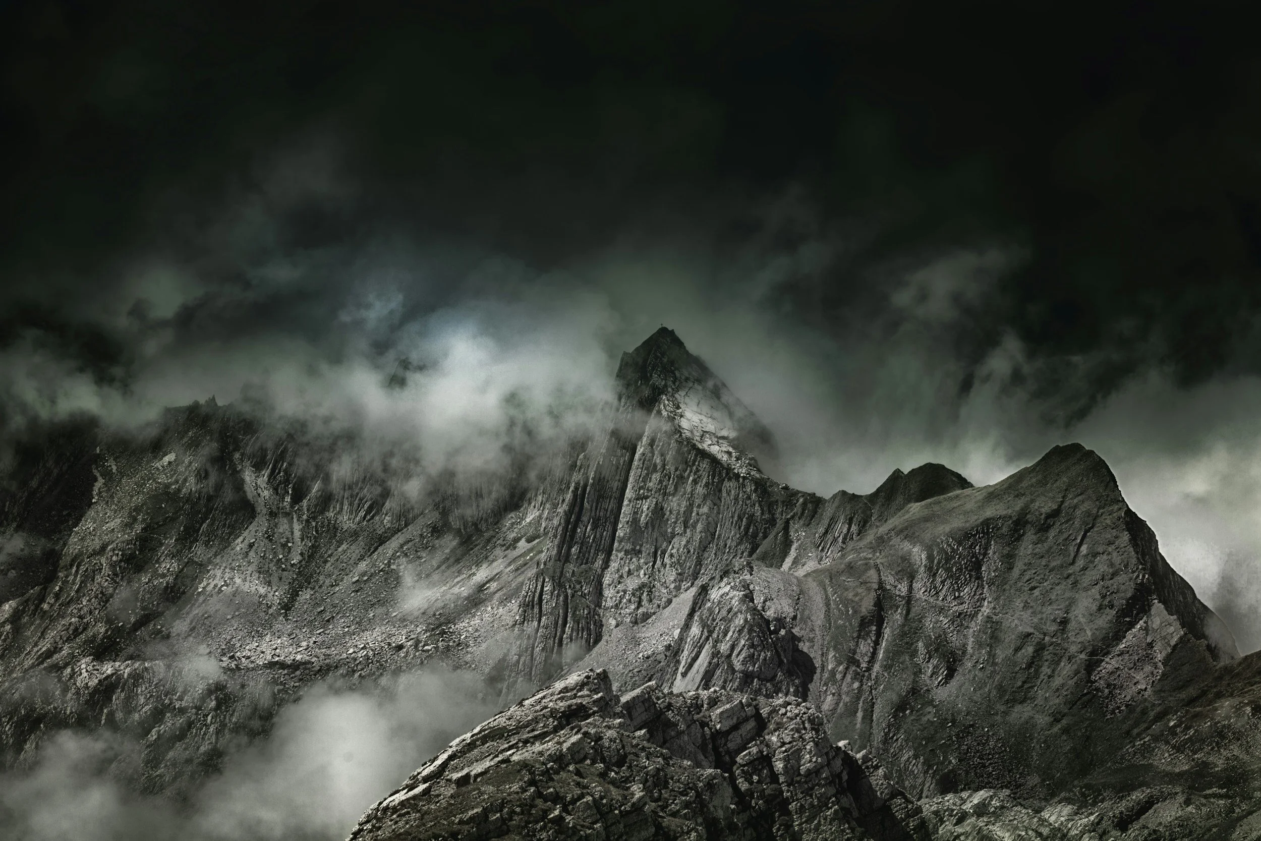 Dark, cloudy mountain landscape with steep rock formations and mist or clouds surrounding the peaks.