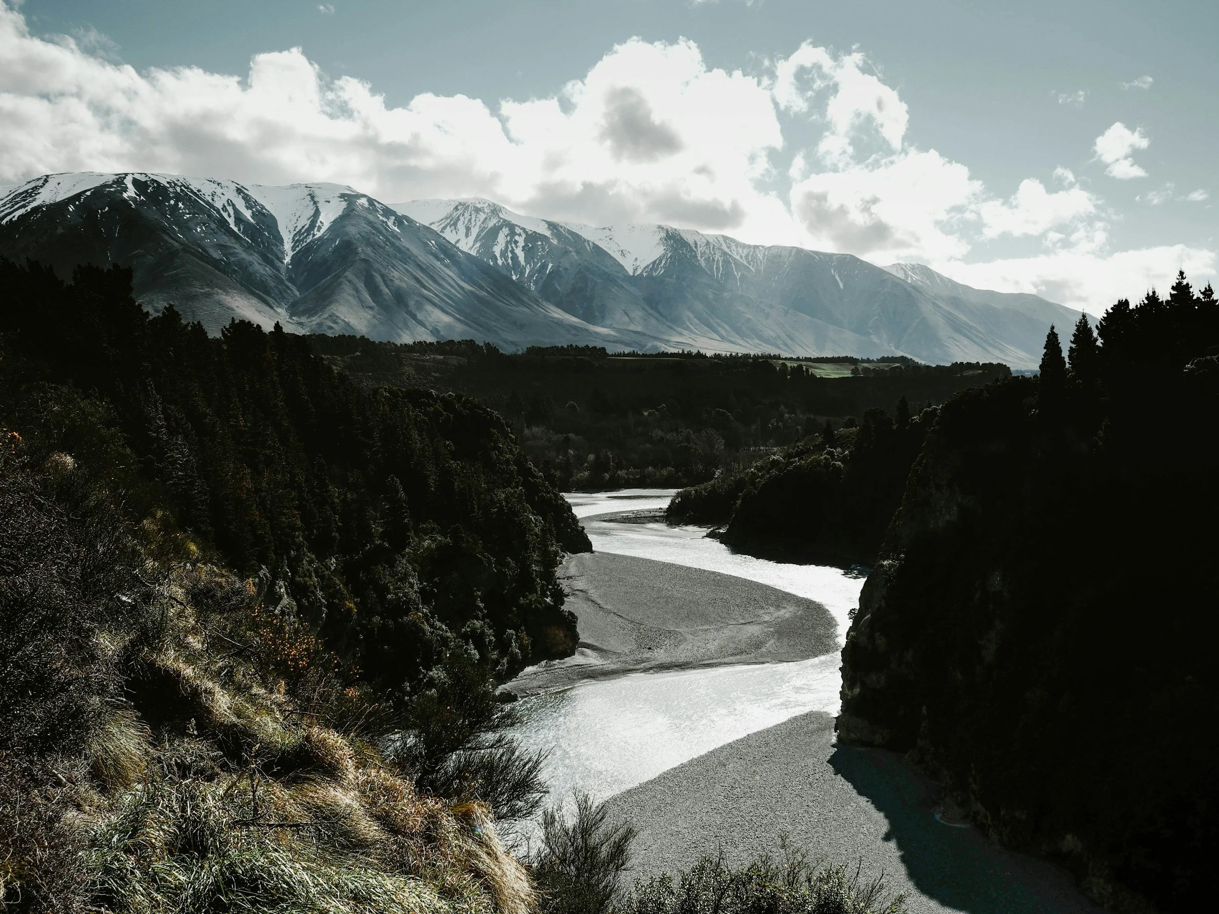 A mountain landscape with snow-capped peaks, a winding river, and dense forested hills under a partly cloudy sky.