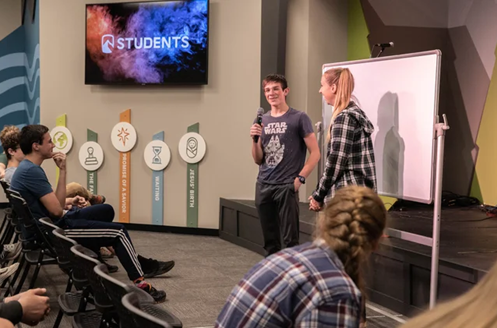 A young man in a gray Star Wars T-shirt holding a microphone stands next to a young woman in a black and white plaid shirt during a presentation or speech at a church or community center. Several people are seated, listening attentively. A large screen with the word 'STUDENTS' displayed is visible behind them. The room has colorful wall decorations with icons and labels, including the phrase 'PROMISE OF A SAVIOR' and 'HIS BISHER.'