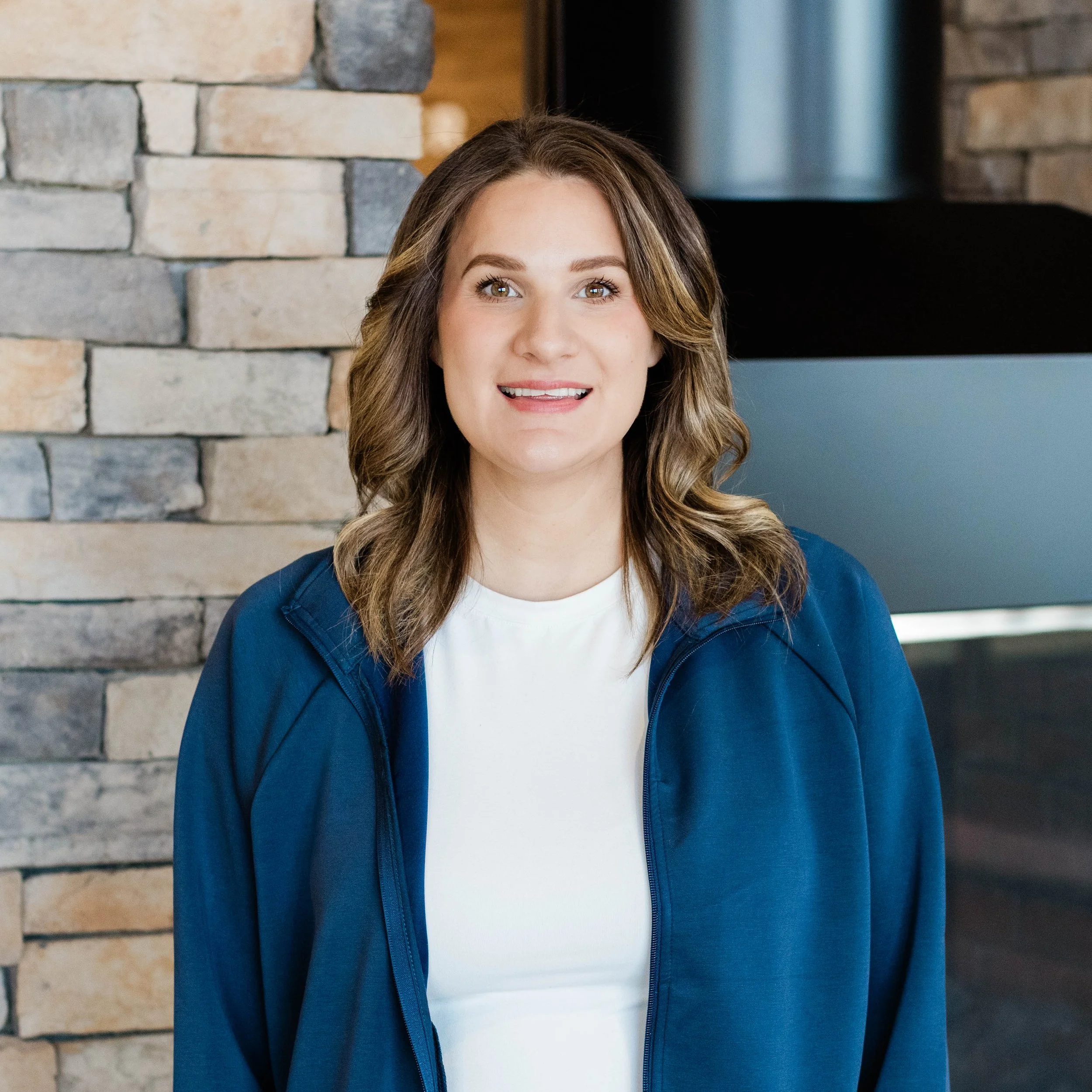 A smiling woman with shoulder-length wavy brown hair, wearing a black top, standing in front of a stone fireplace with a modern design.