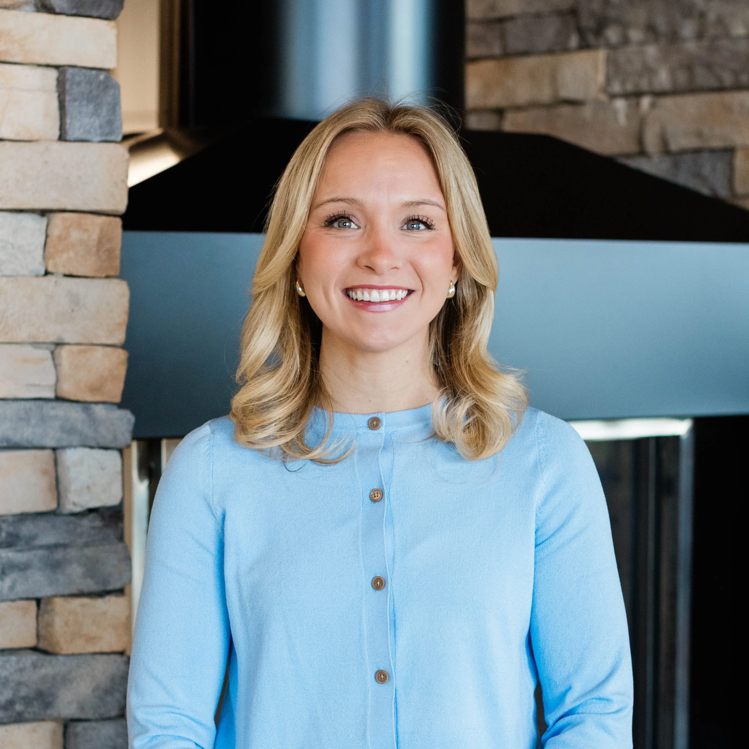 A woman with shoulder-length blonde hair smiling in front of a stone fireplace.