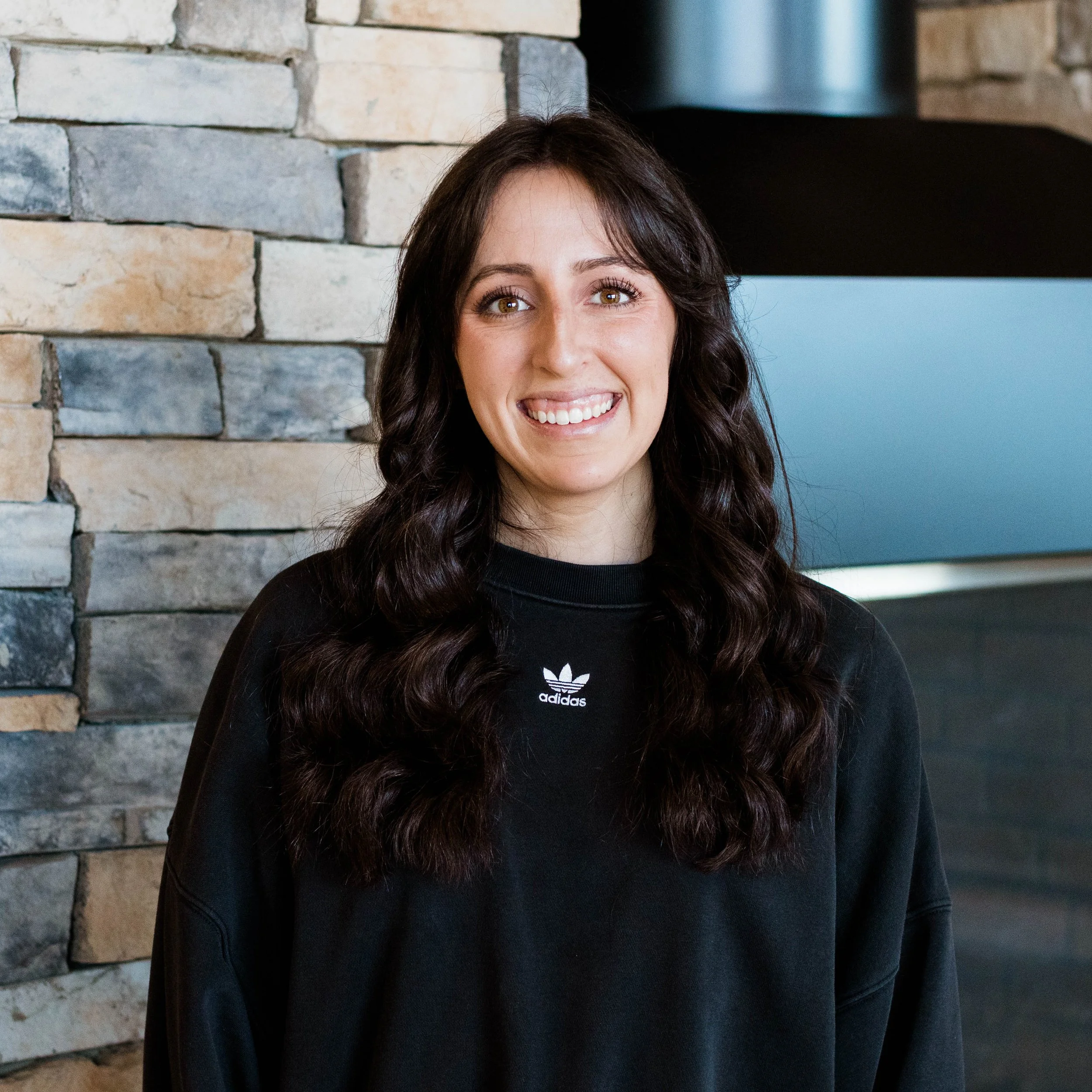 A woman with long, curly dark hair smiling in front of a stone fireplace.