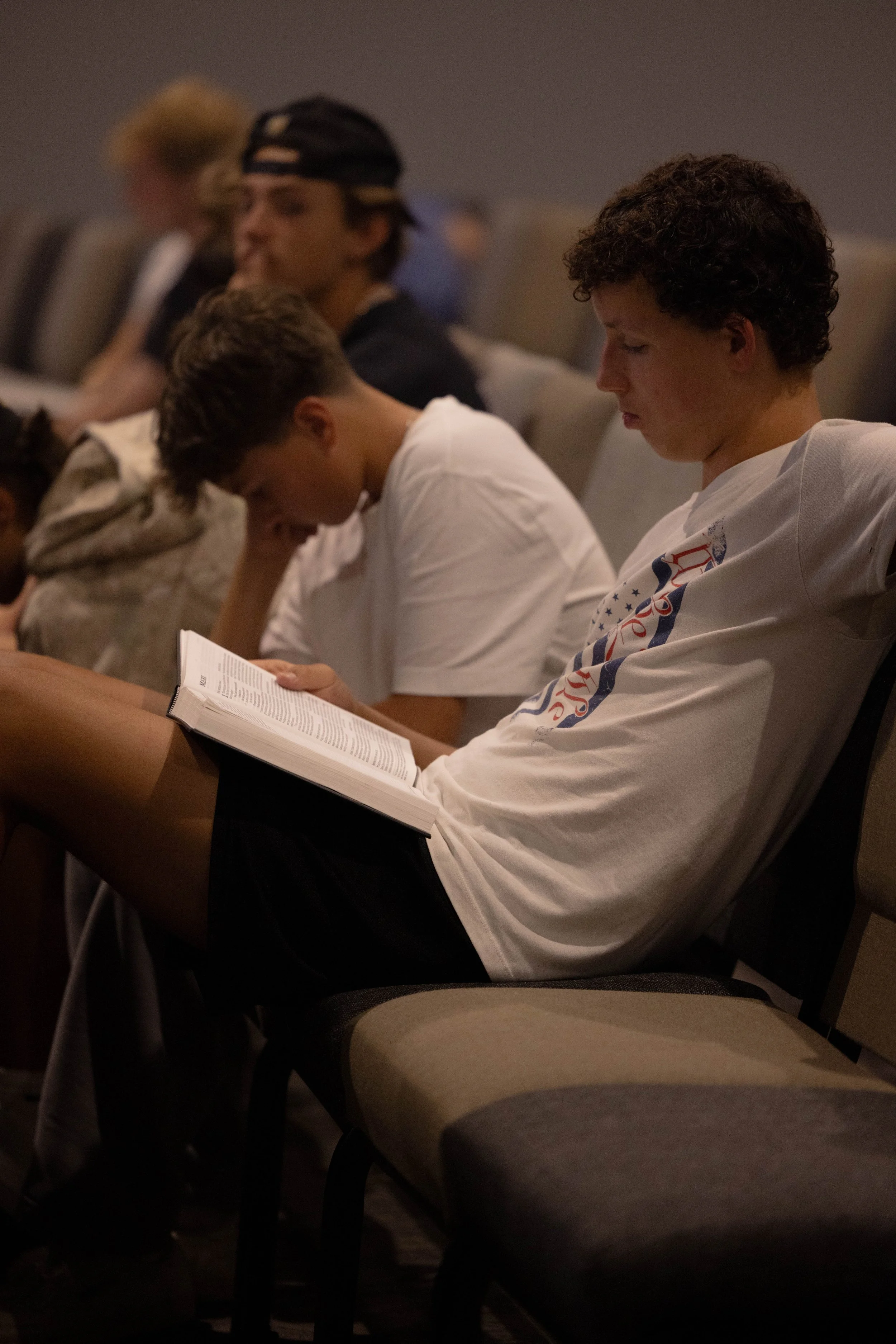 A group of people, mostly young adults, standing closely together with their arms around each other's shoulders in prayer or reflection, facing towards the front of the room where two individuals are standing at a distance.