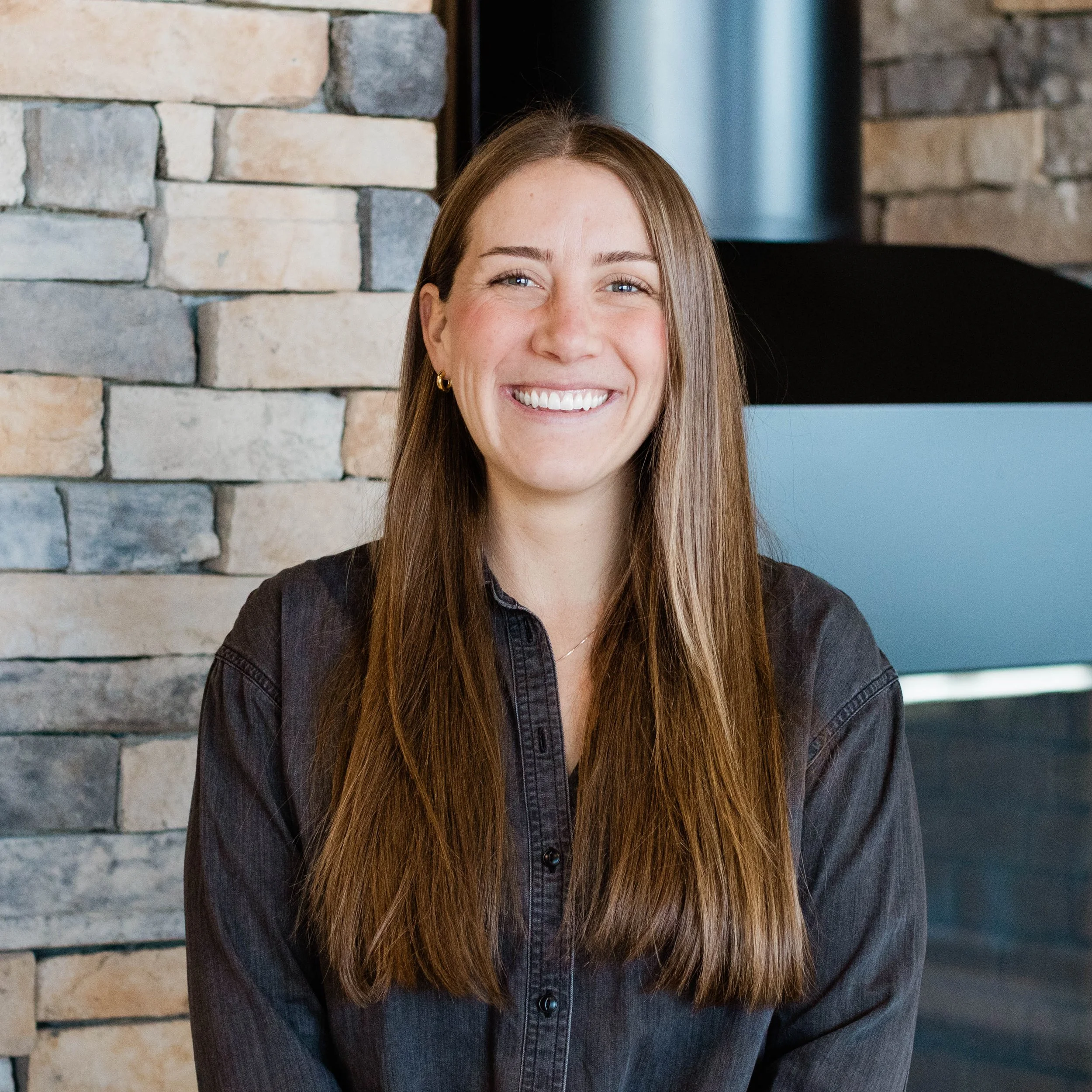 A woman with long brown hair, wearing a green t-shirt, smiling in front of a stone fireplace.