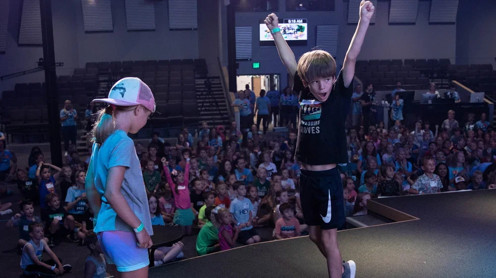 A young boy with arms raised in celebration and a girl with a cap holding a tablet on stage, with a large audience in the background.