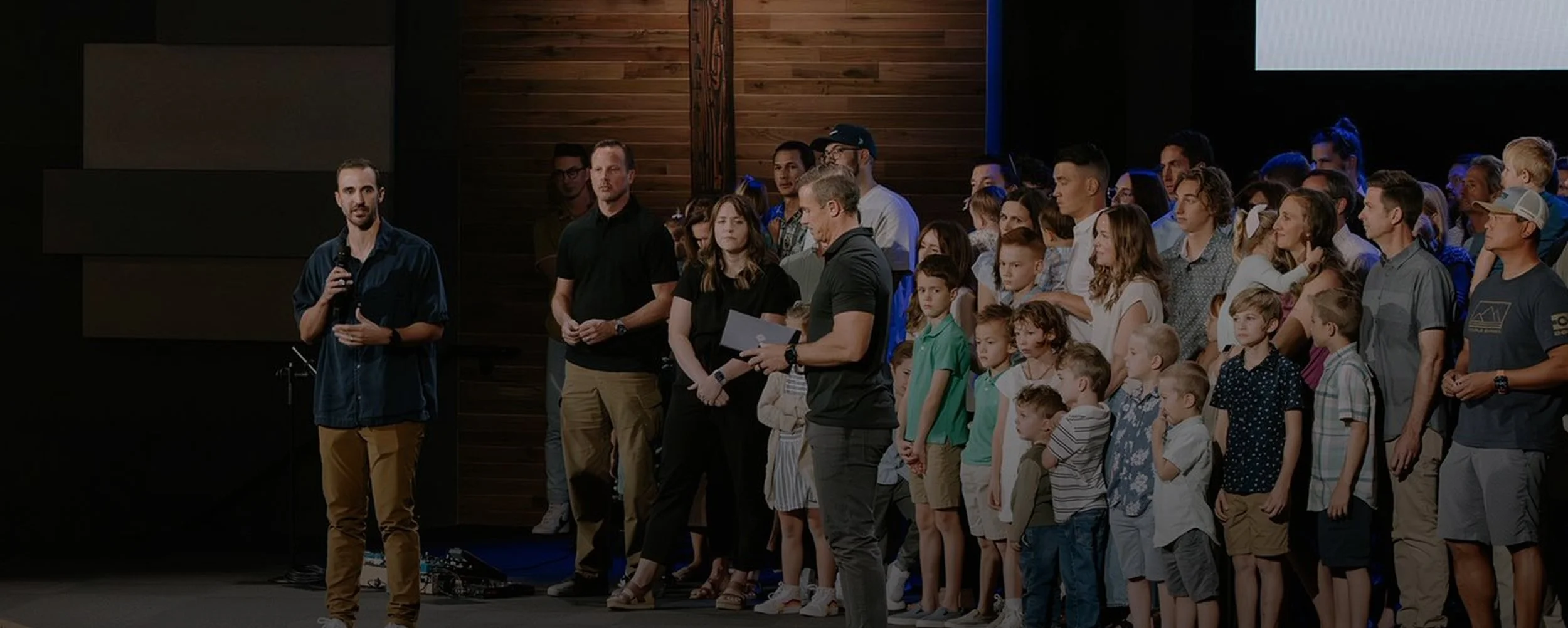 A man in a blue shirt and tan pants holding a microphone speaking to a group of people, including adults and children, on a stage with a wooden wall background.