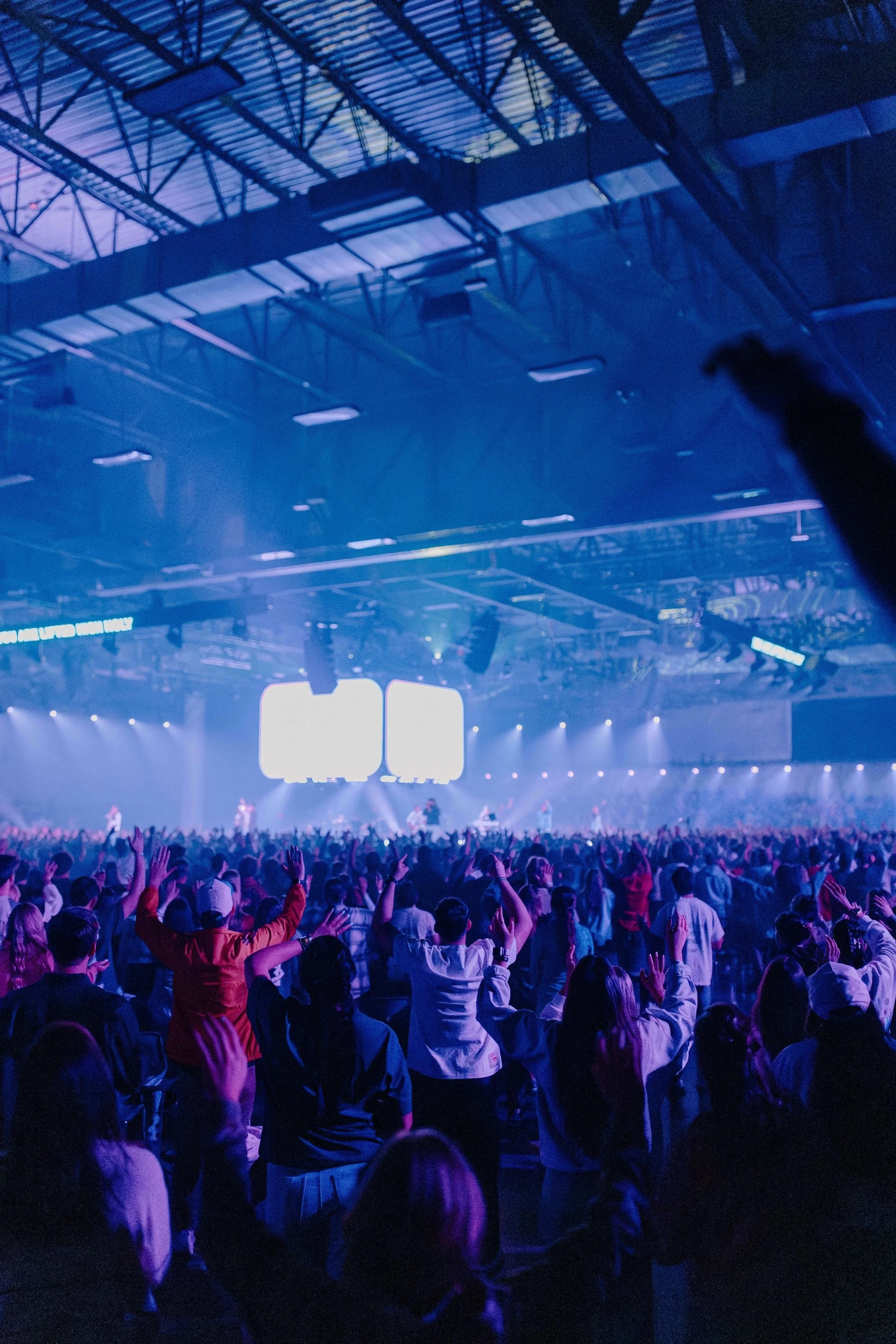 Crowd of people at a concert or event in a large indoor arena with a stage, lighting, and screens, enjoying the show.