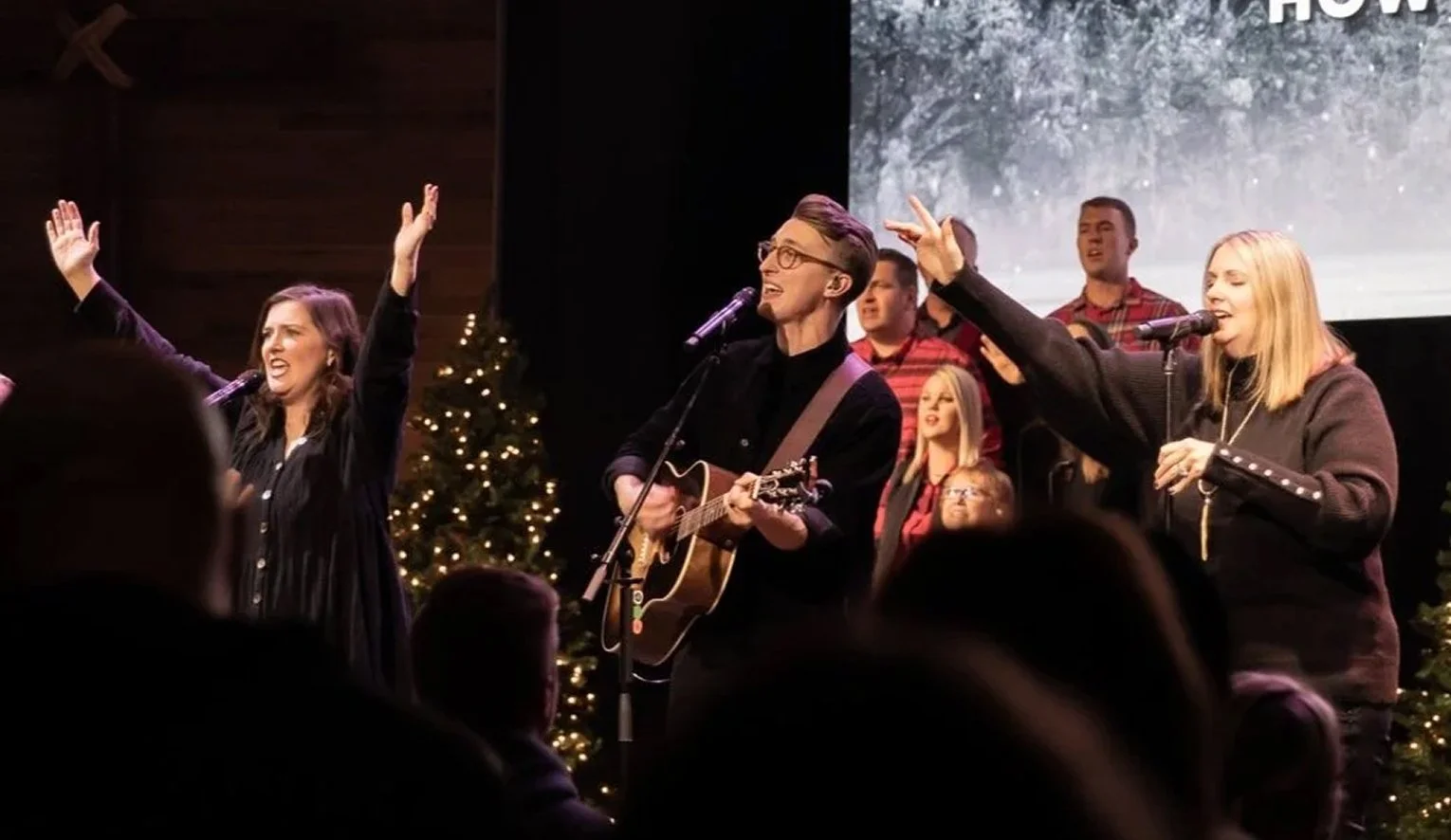 People singing and playing guitar on stage during a Christmas concert, decorated with Christmas trees.