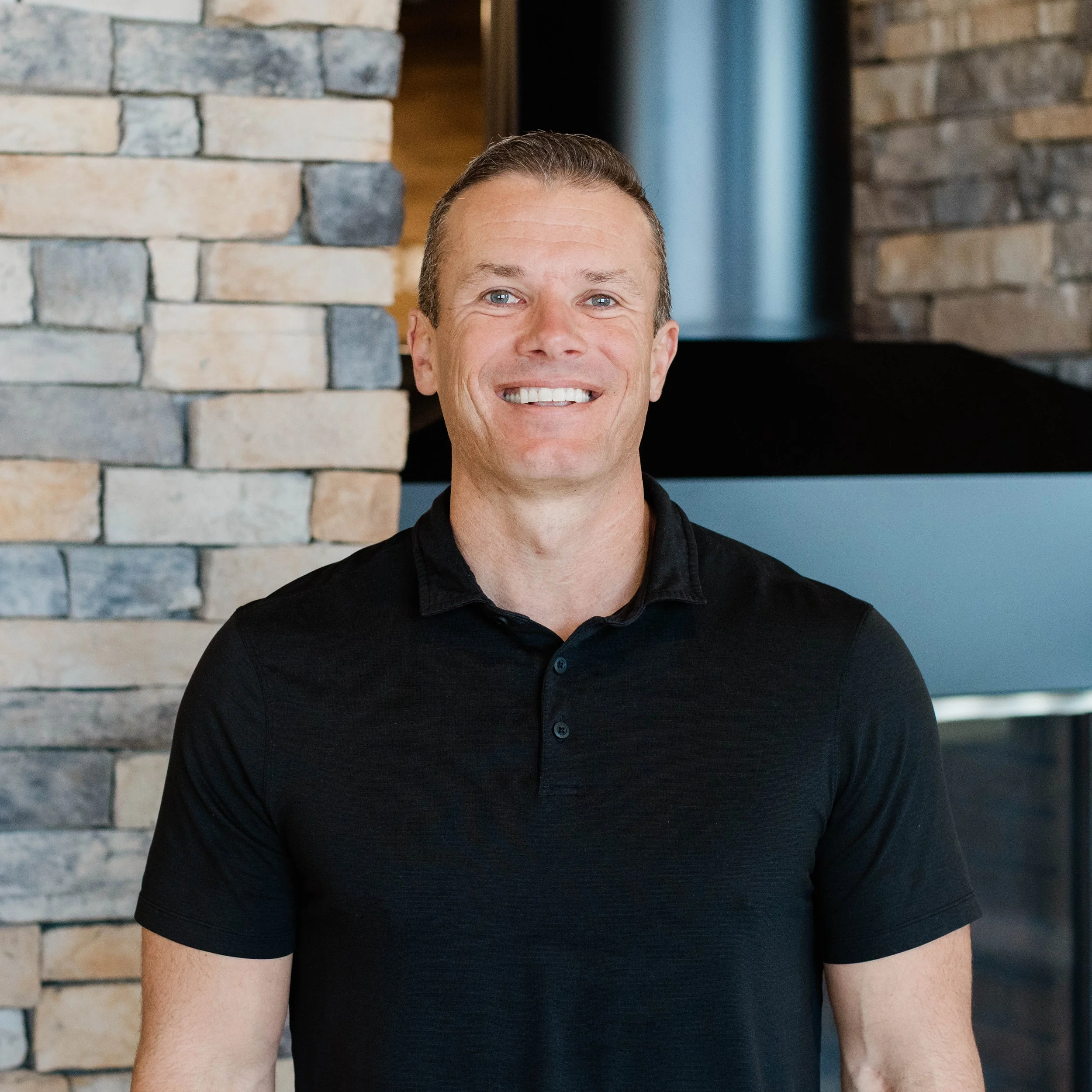 Smiling man in a black polo shirt standing in front of a stone wall with a fireplace.