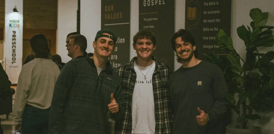 Three young men smiling and giving thumbs up in a church lobby with personal belief signs and a plant in the background.