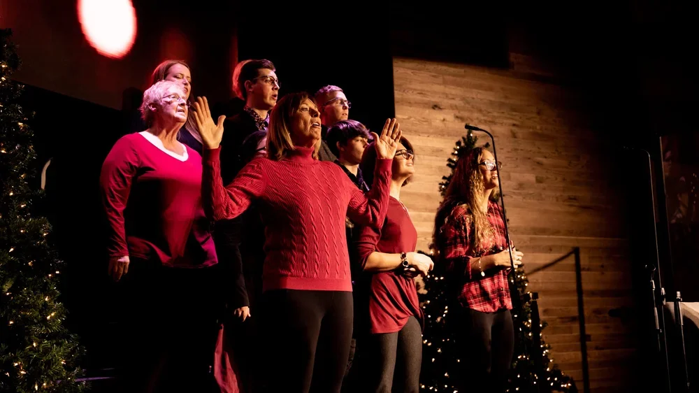 Group of women singing during a Christmas performance, dressed in red and holiday-themed clothing, standing on stage decorated with Christmas trees.