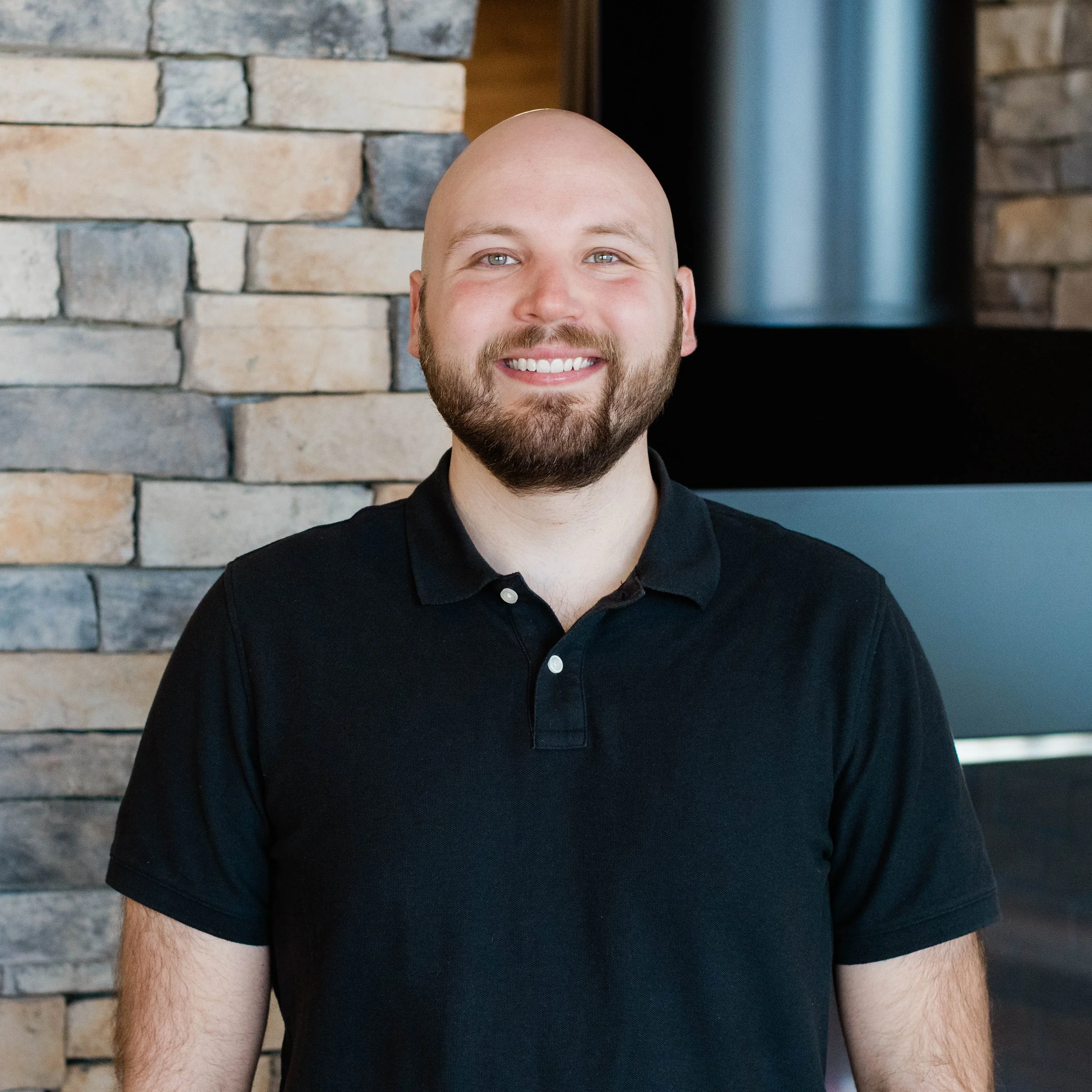 A smiling man with a beard and short hair, wearing a dark gray polo shirt, standing indoors near a stone fireplace.