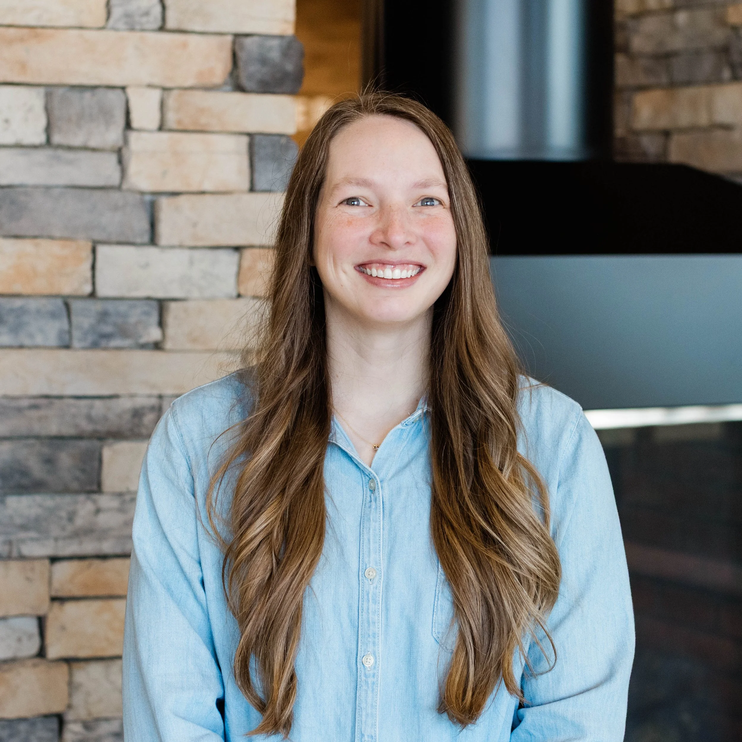 A young woman with long wavy hair smiling, wearing a black t-shirt, standing in front of a stone wall.