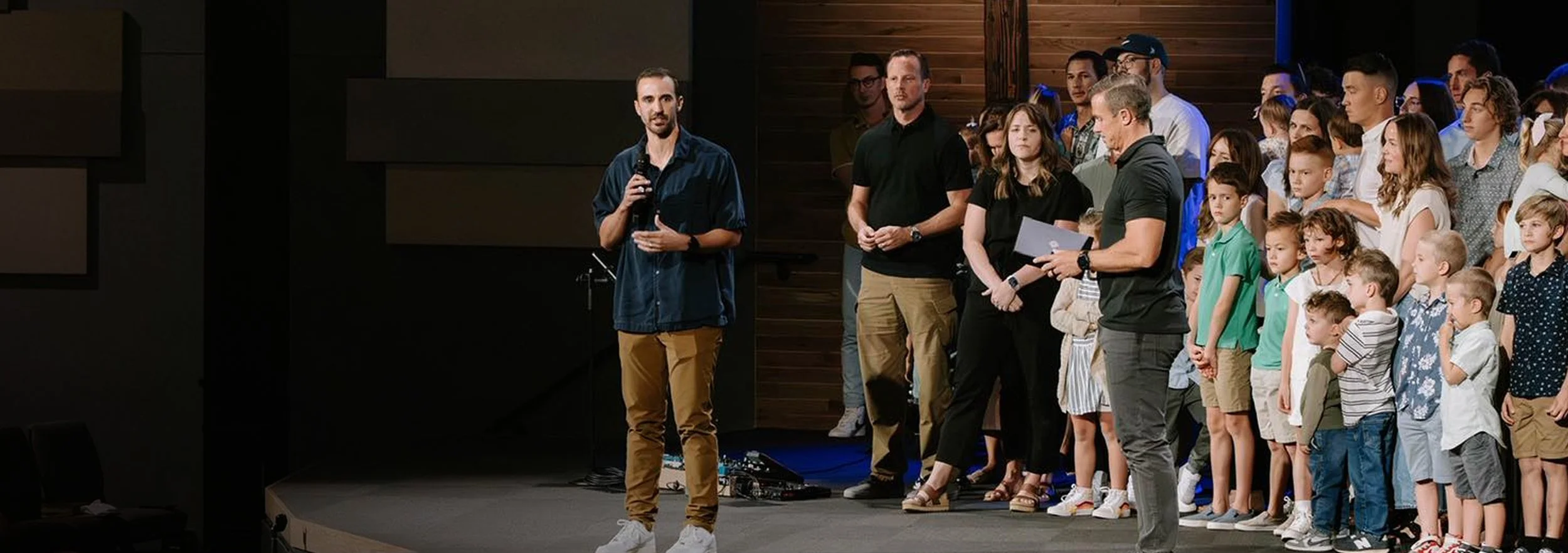 A man in a dark shirt and beige pants speaks into a microphone on a stage, with a large group consisting of children and adults standing behind him in a room with dark wood paneling.