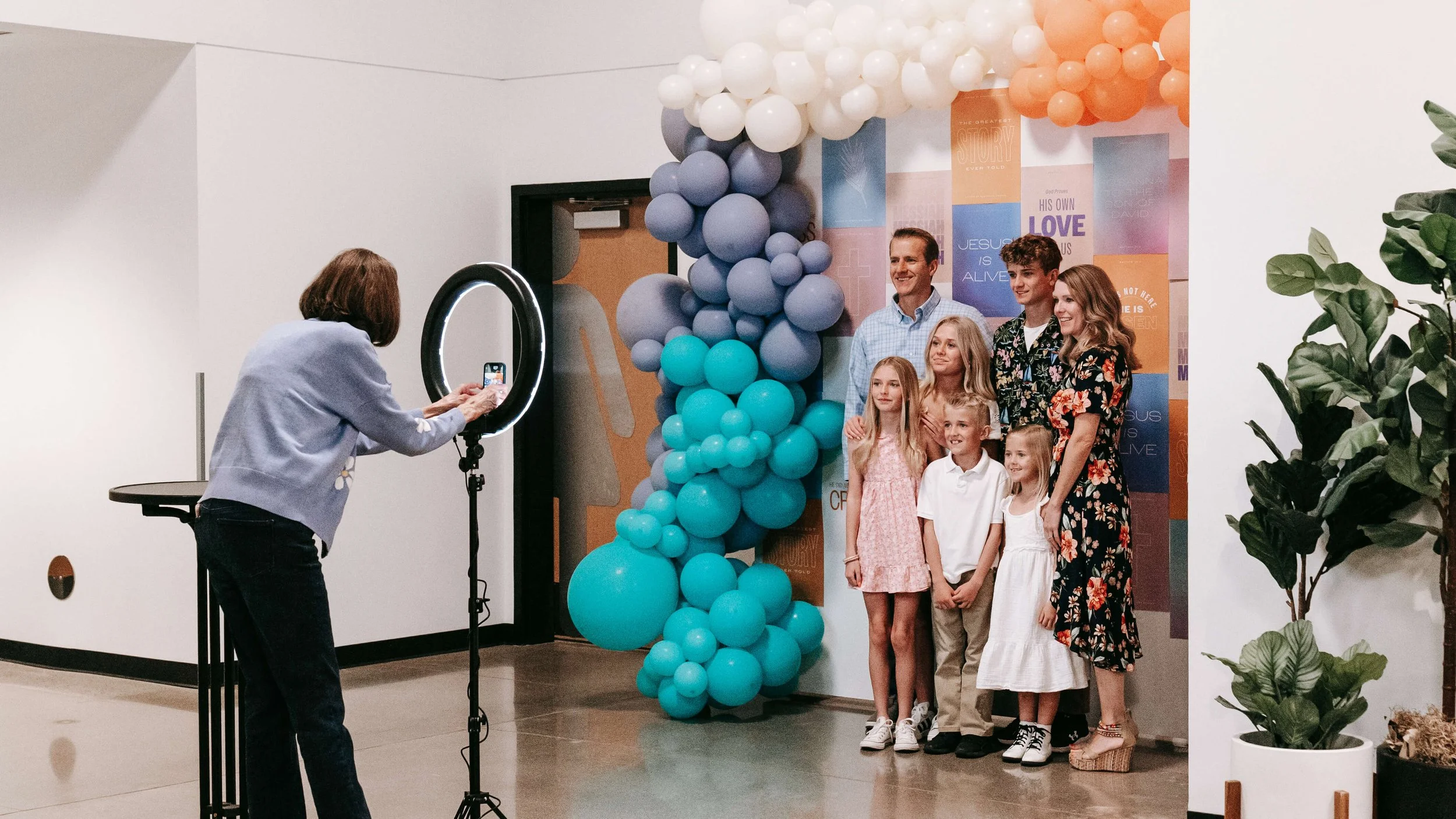 A woman takes a photo of a family posing in front of a balloon decoration with a ring light. The family has two adults and five children, standing in front of a colorful backdrop with various messages, in a well-lit indoor space with a potted plant on the right.