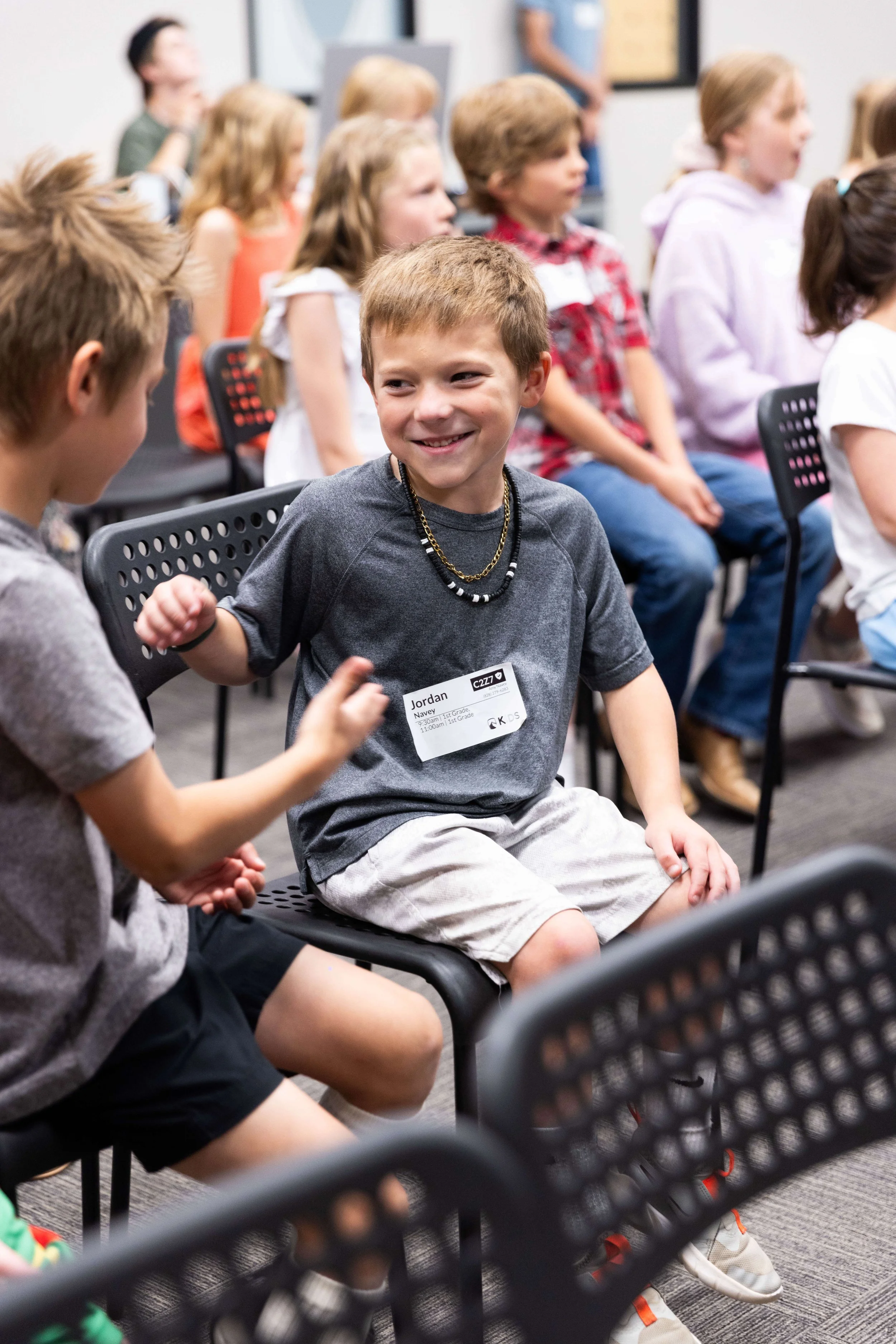 A group of children sitting on chairs in a classroom or event setting, smiling and engaging with each other. Focus on a boy in a gray shirt with a name tag that reads 'Jordan,' talking to another child.