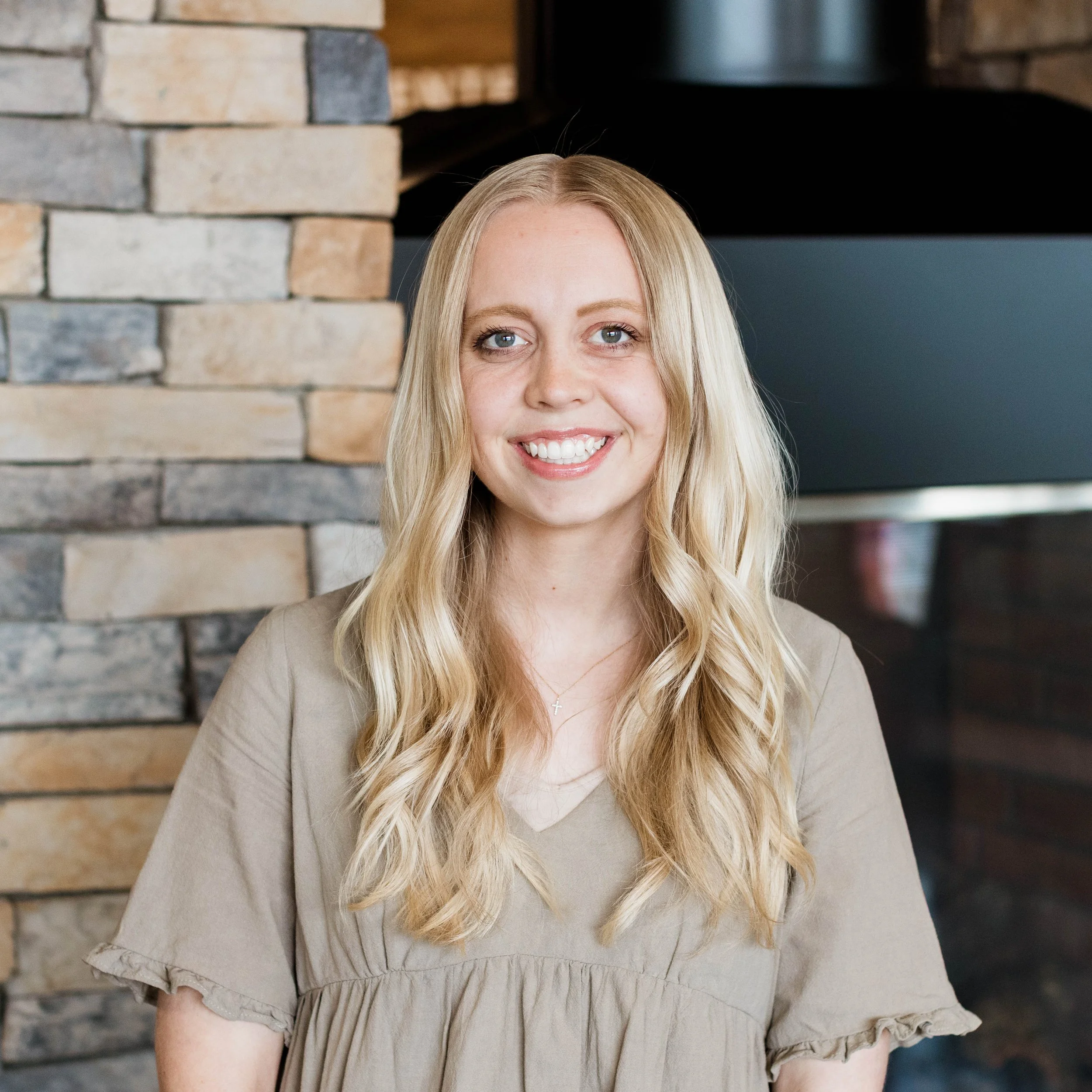 A young woman with long, wavy blonde hair smiling indoors, standing in front of a stone fireplace.