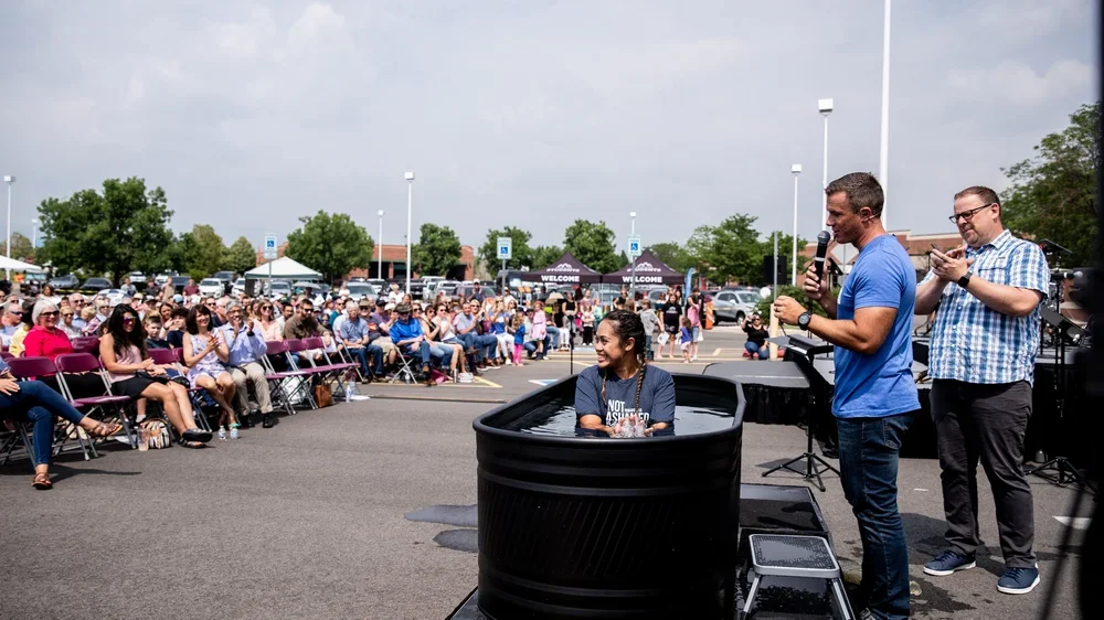 Woman in baptismal tank at outdoor event, man with microphone and another man clapping, audience seated in chairs, tents, and trees in background.