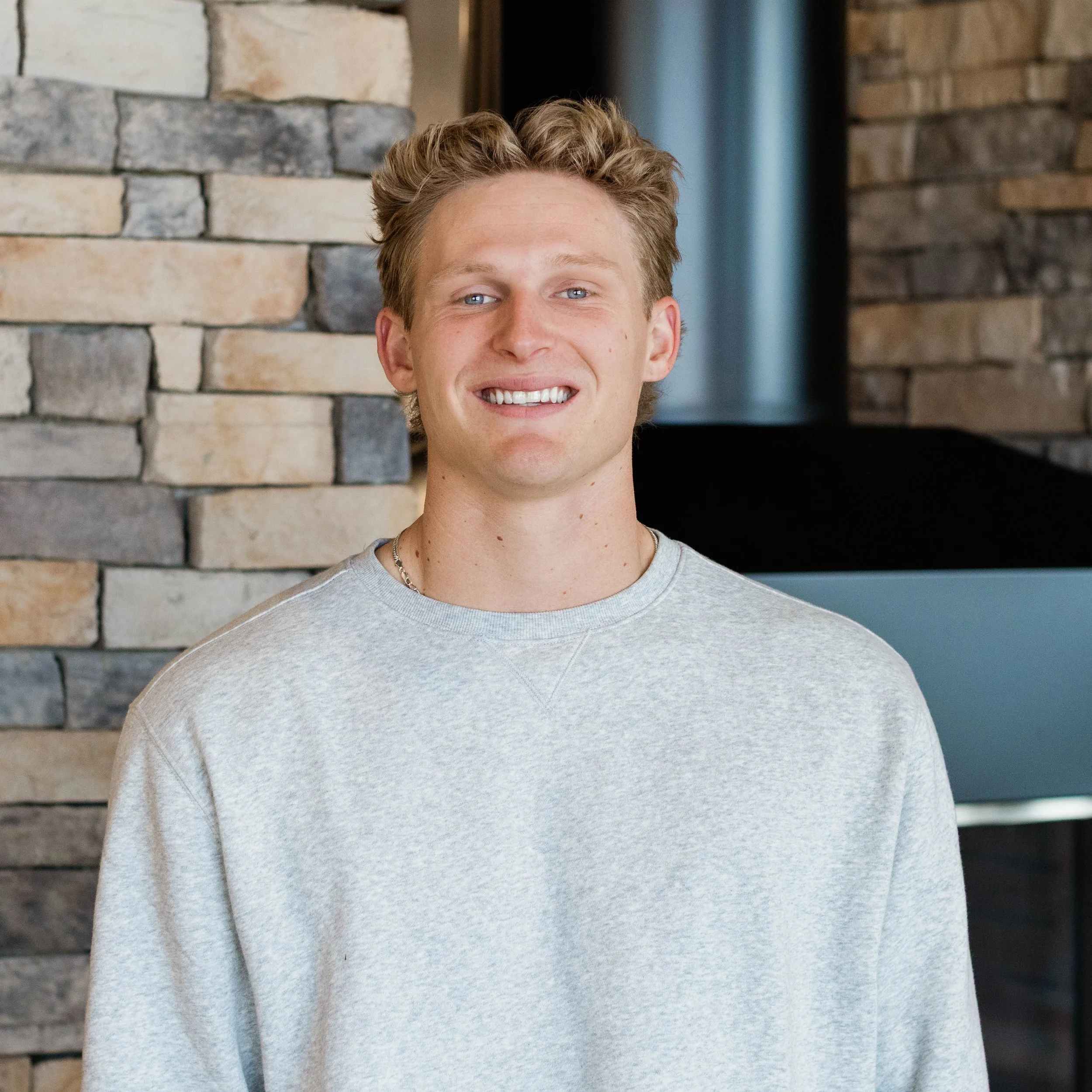 A young man with curly red hair and blue eyes, smiling, wearing a light gray T-shirt, standing indoors near a brick wall.