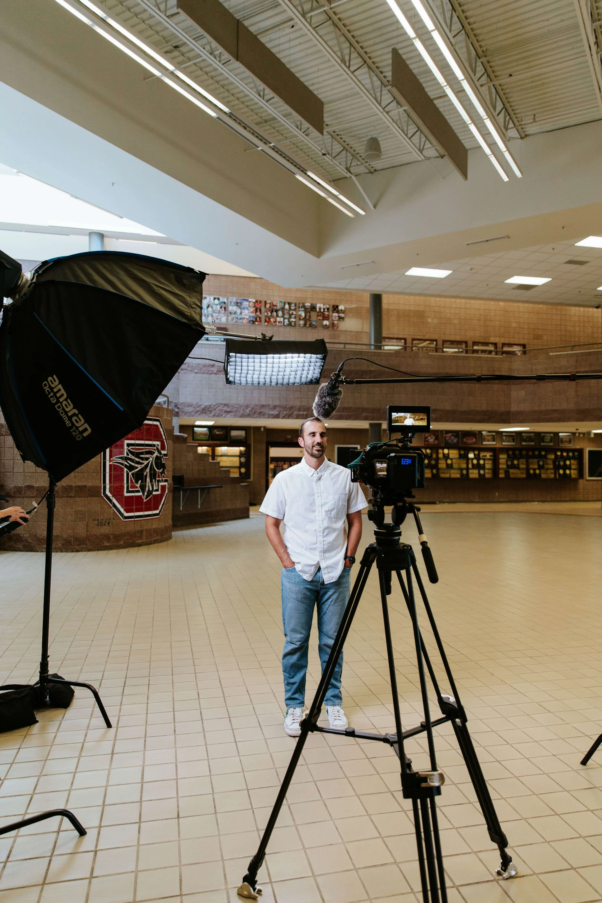 A man in a white shirt and jeans being filmed in a large indoor space with professional lighting and camera equipment, possibly a school or public building lobby.