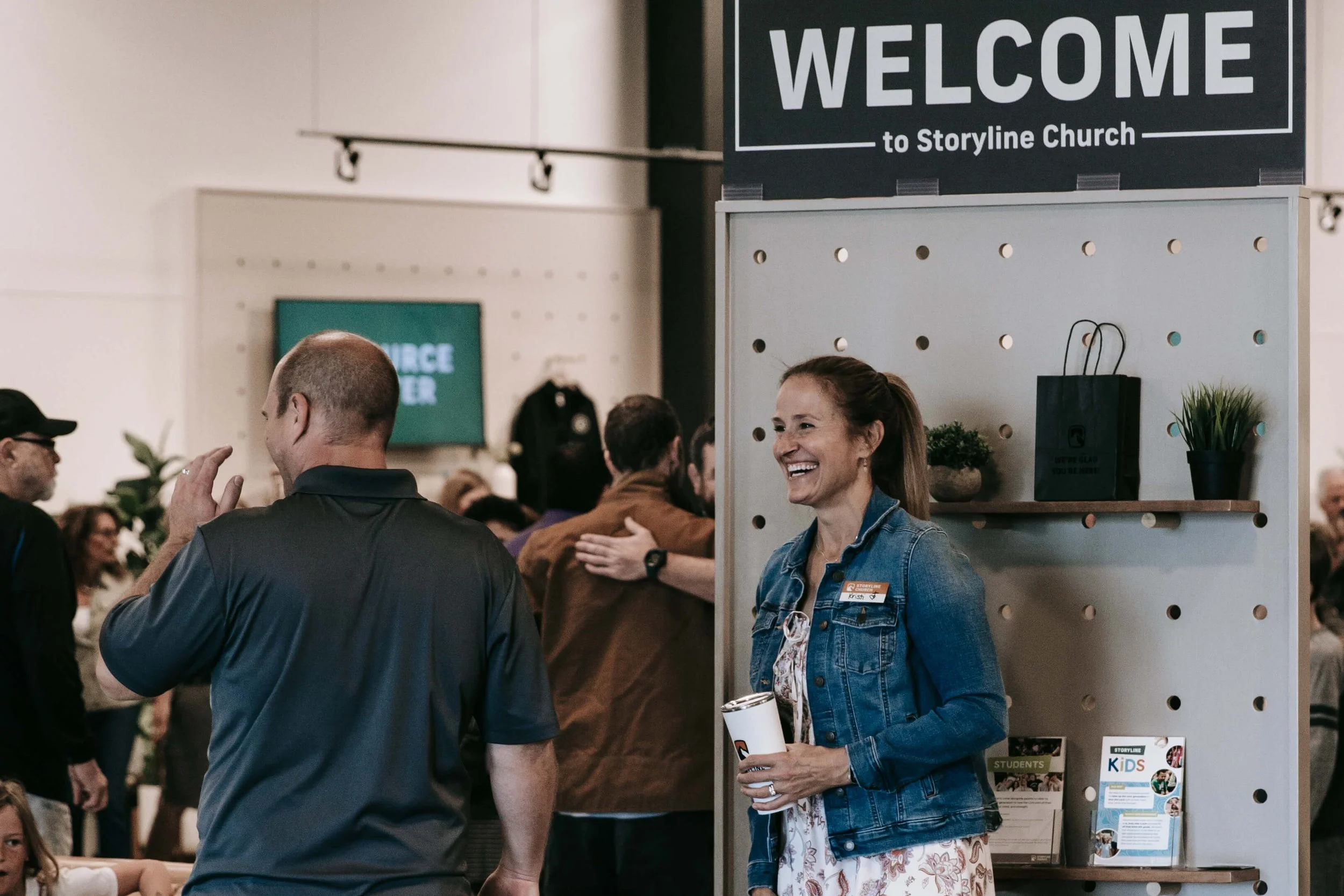 Smiling woman with a denim jacket talking to a man at a church welcome event, with other attendees visible in the background.