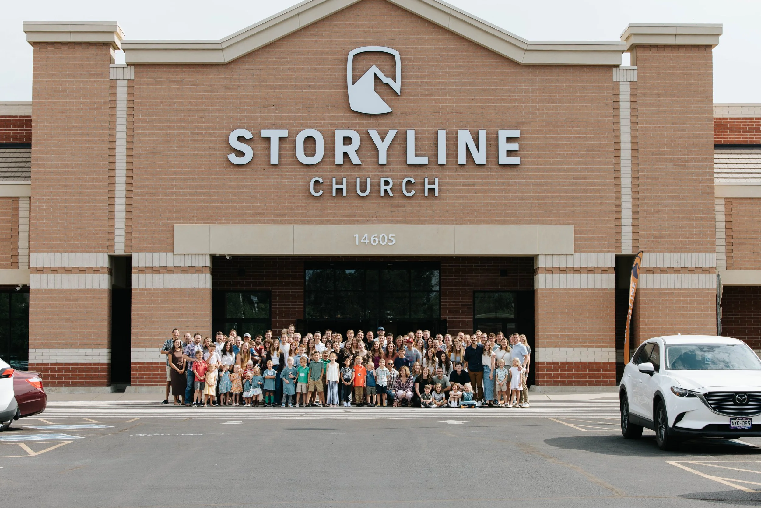 A large group of people, including children and adults, standing in front of a brick church building with a sign that reads 'Storyline Church' and the number '14605' above the entrance.