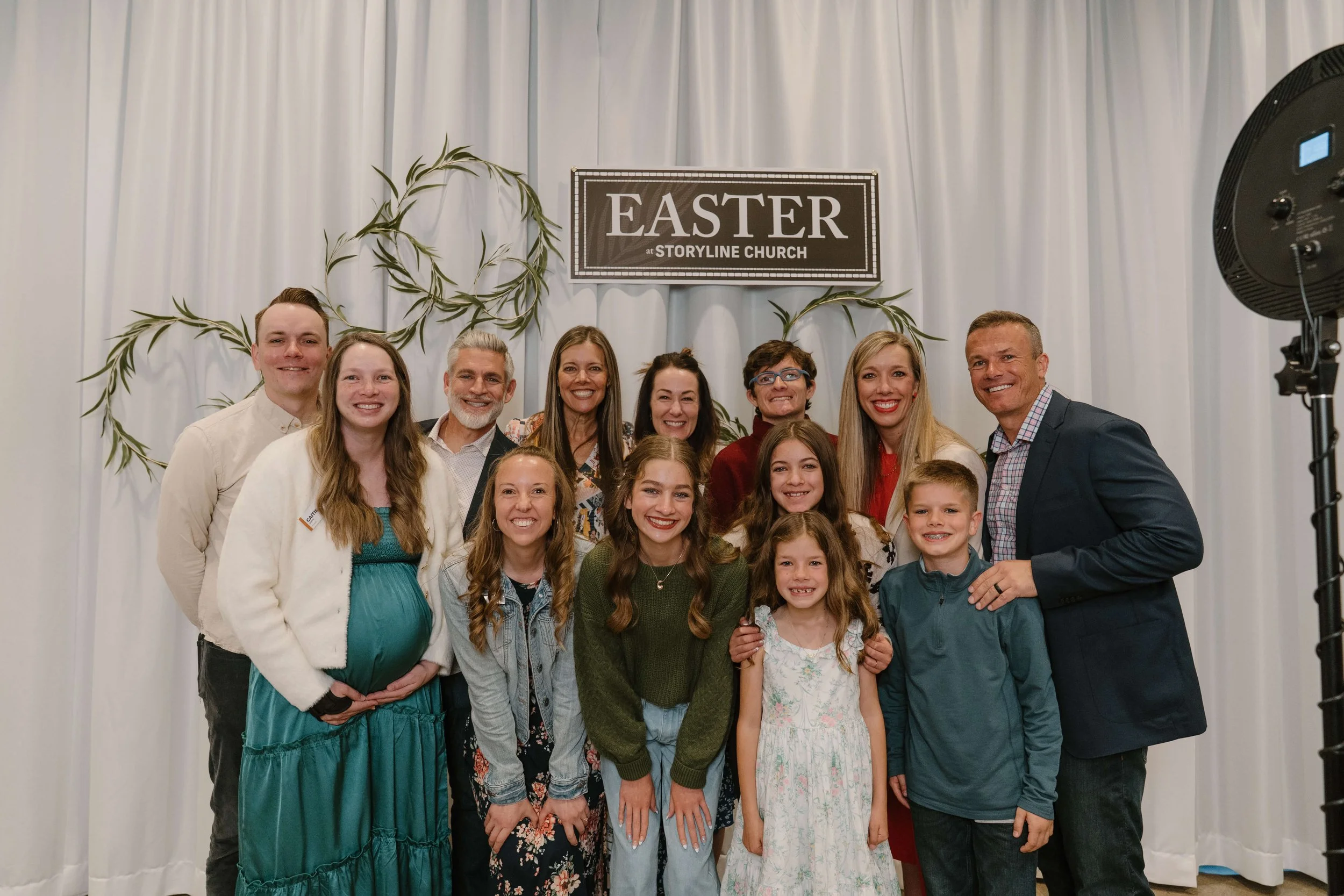 Group of people gathered for Easter celebration at Storyline Church, smiling and posing for a photo in front of a white backdrop with a sign that reads 'EASTER at STORYLINE CHURCH' and decorative greenery.