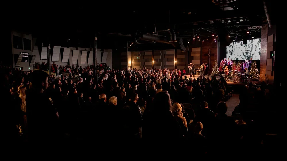 Large audience attending a Christmas-themed concert with musicians on stage, decorated with Christmas trees and lights.
