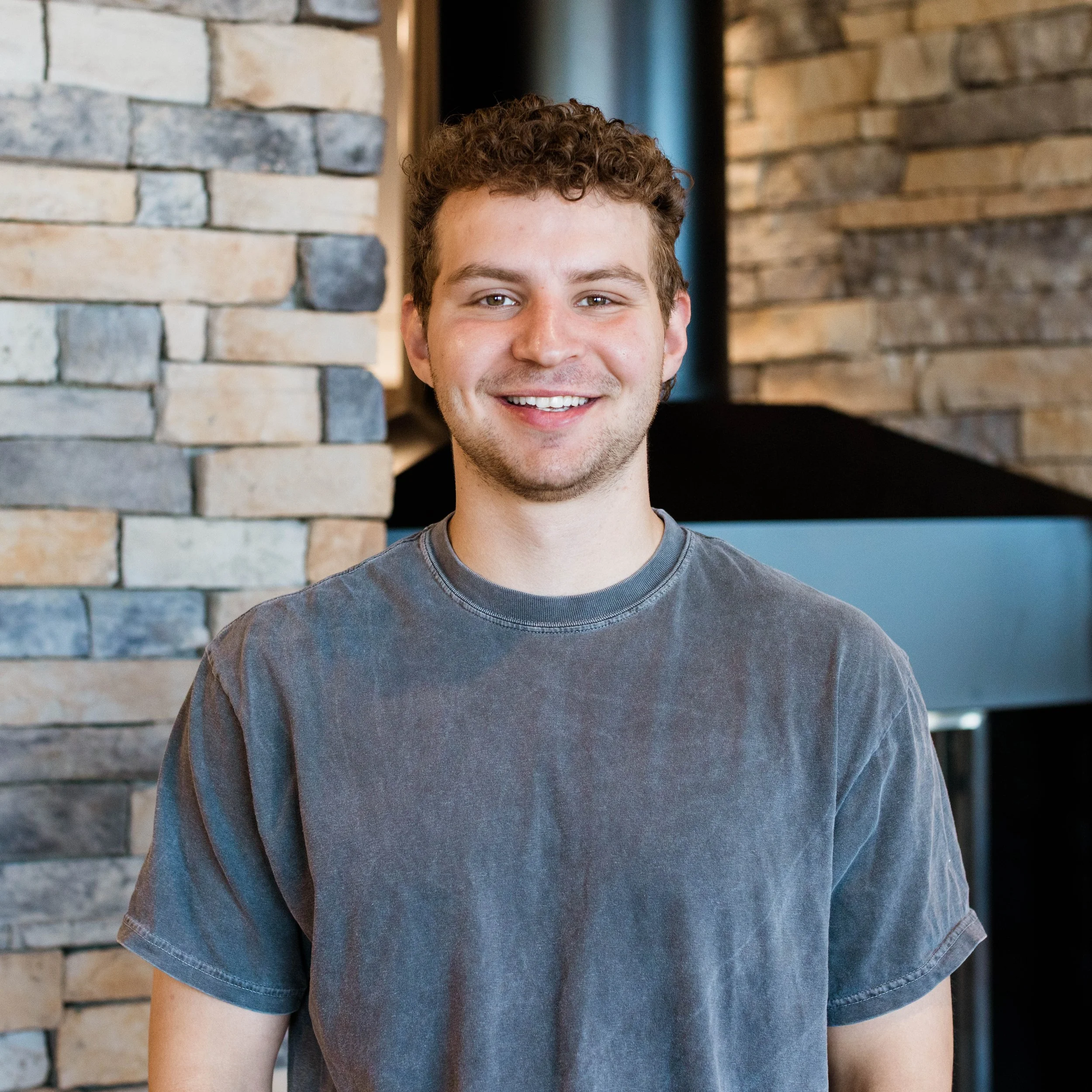 A smiling young man with curly brown hair and a mustache, wearing a black Carhartt T-shirt, standing indoors in front of a stone fireplace.