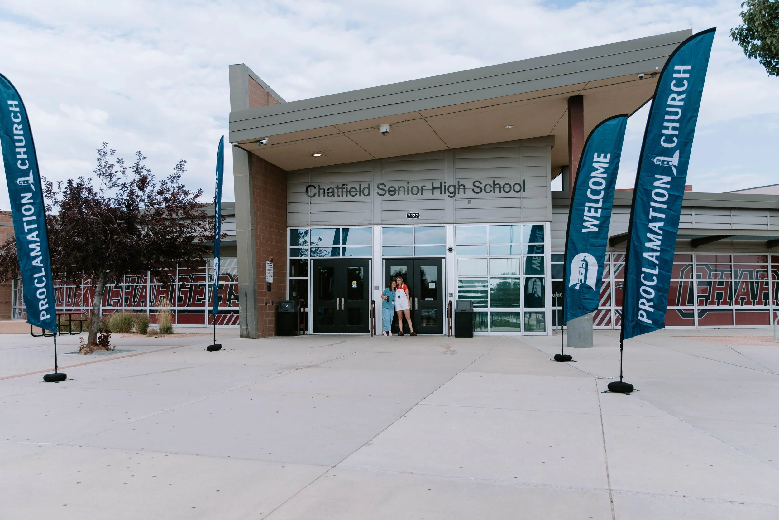 The entrance of Chatfield Senior High School with two people standing in the doorway, surrounded by blue banners that read 'Welcome' and 'Proclamation Church.'