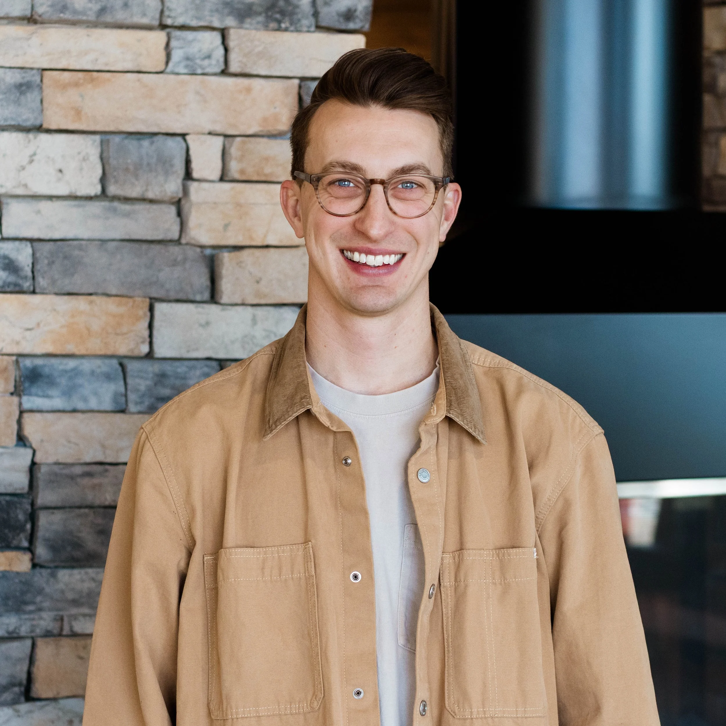 A smiling young man with glasses and short brown hair standing in front of a stone fireplace in a room with wooden furniture.