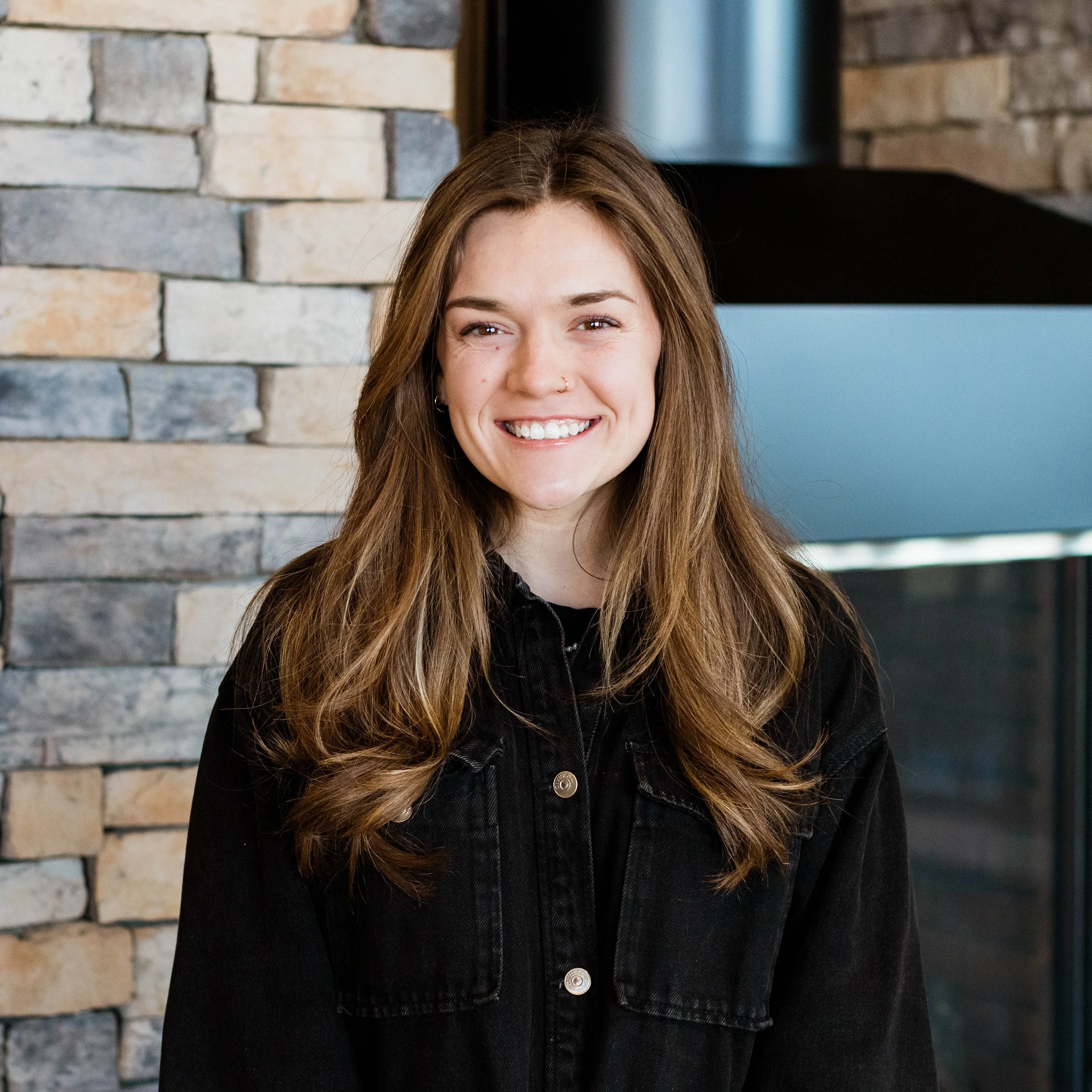 A young woman with long wavy brown hair smiling, wearing a black jacket over a white shirt, standing in front of a stone wall with a fireplace.