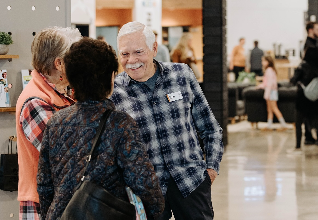 A group of three people, two women and one man, conversing and smiling in a modern indoor setting, with other people and a person in a pink hoodie in the background.