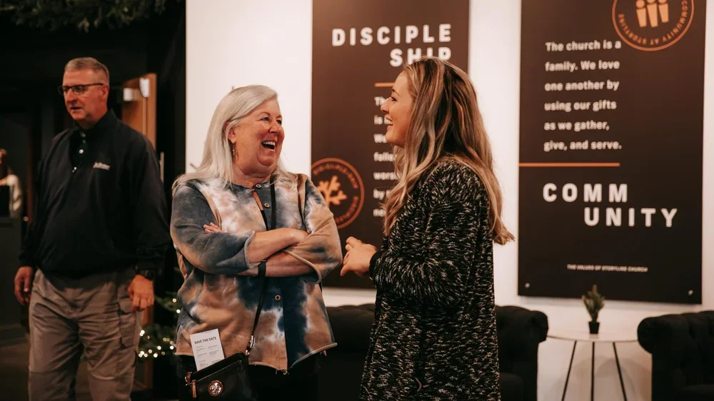 Three people in a church lobby, two women talking and smiling, while a man stands near them. Background features banners about church values and a small potted plant on a table.