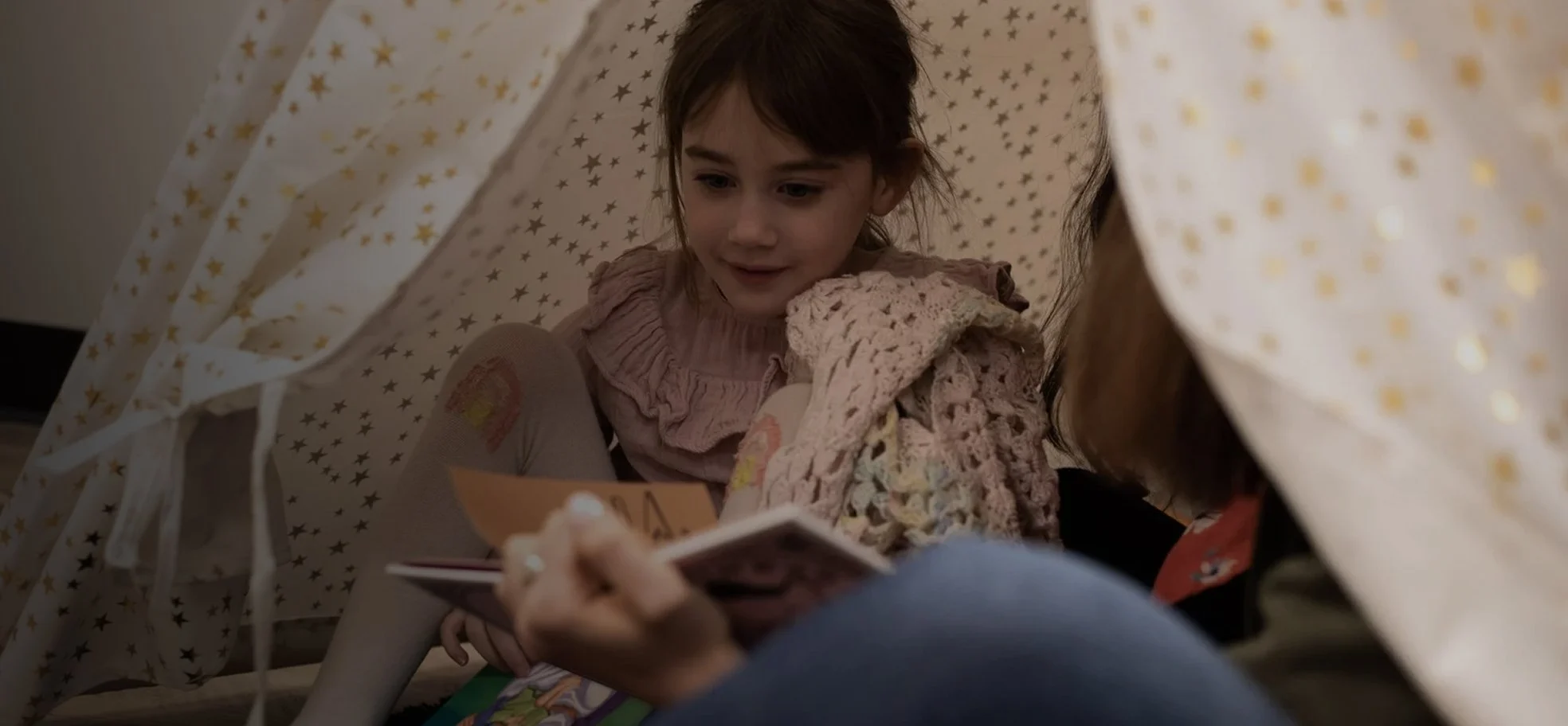 A young girl with red hair and a pink ruffled shirt looks inside a paper card or letter inside a makeshift blanket fort, with star-patterned cloth draped overhead.