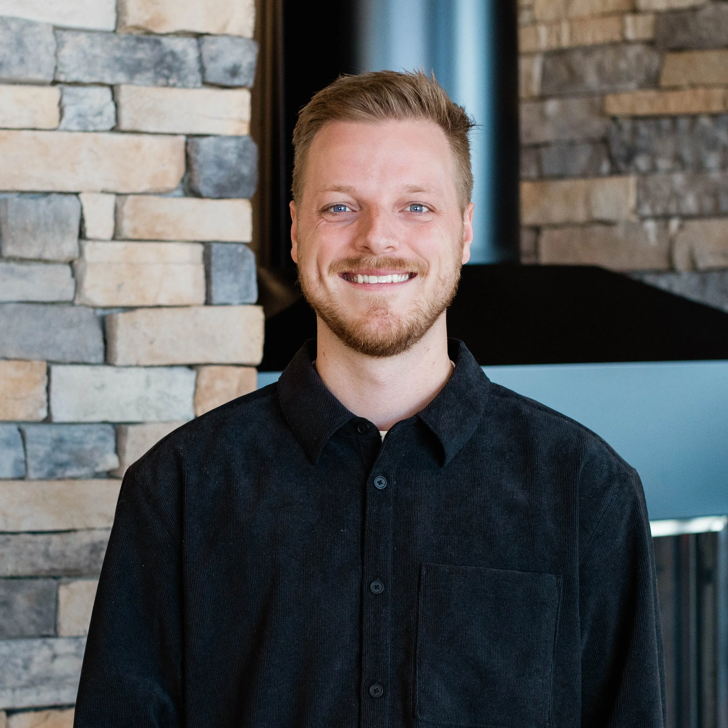 A smiling man with short brown hair and a beard, wearing a dark green shirt, standing in front of a stone brick wall.