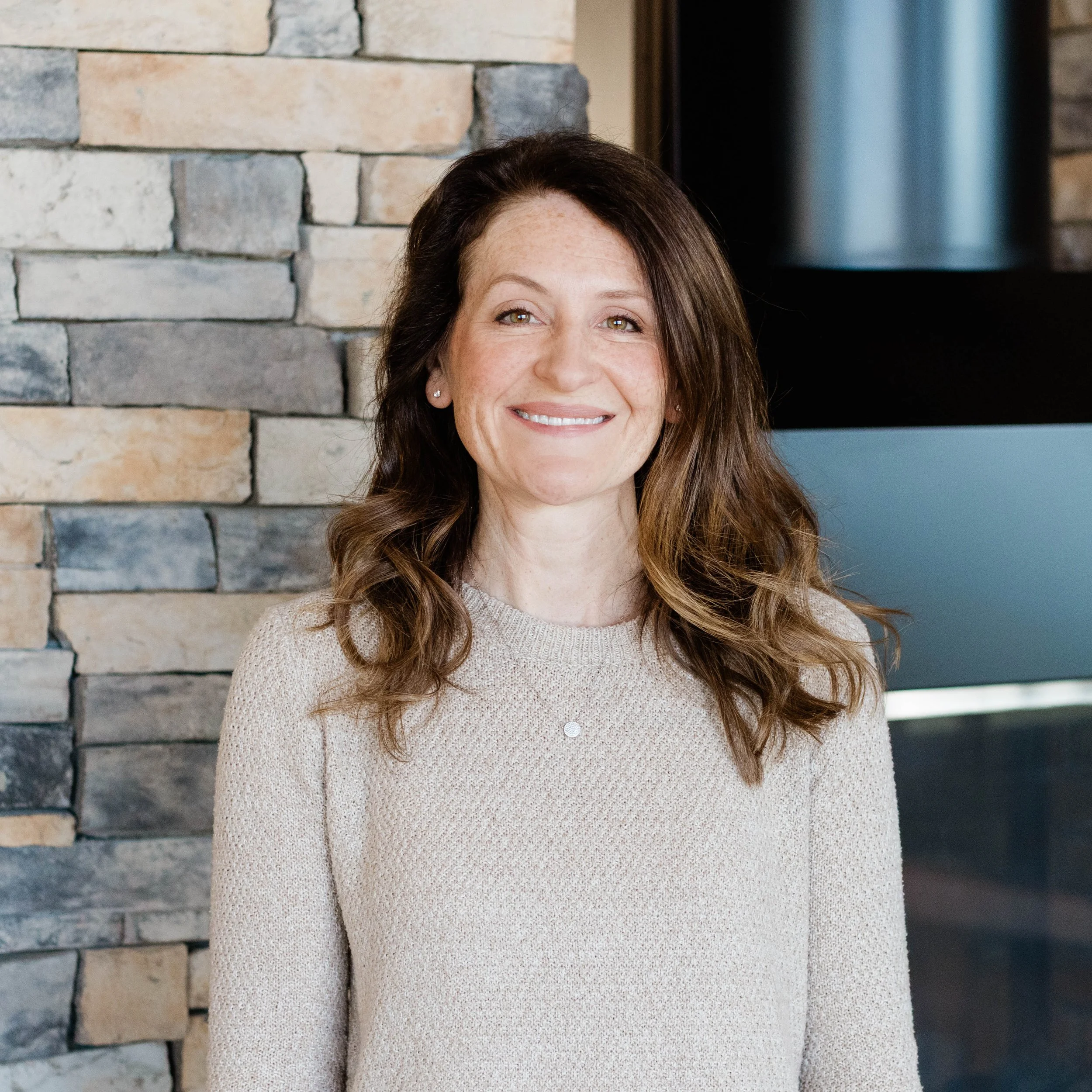A smiling woman with shoulder-length brown hair wearing a red floral-patterned blouse and a long necklace. She is standing in front of a brick wall, indoors.