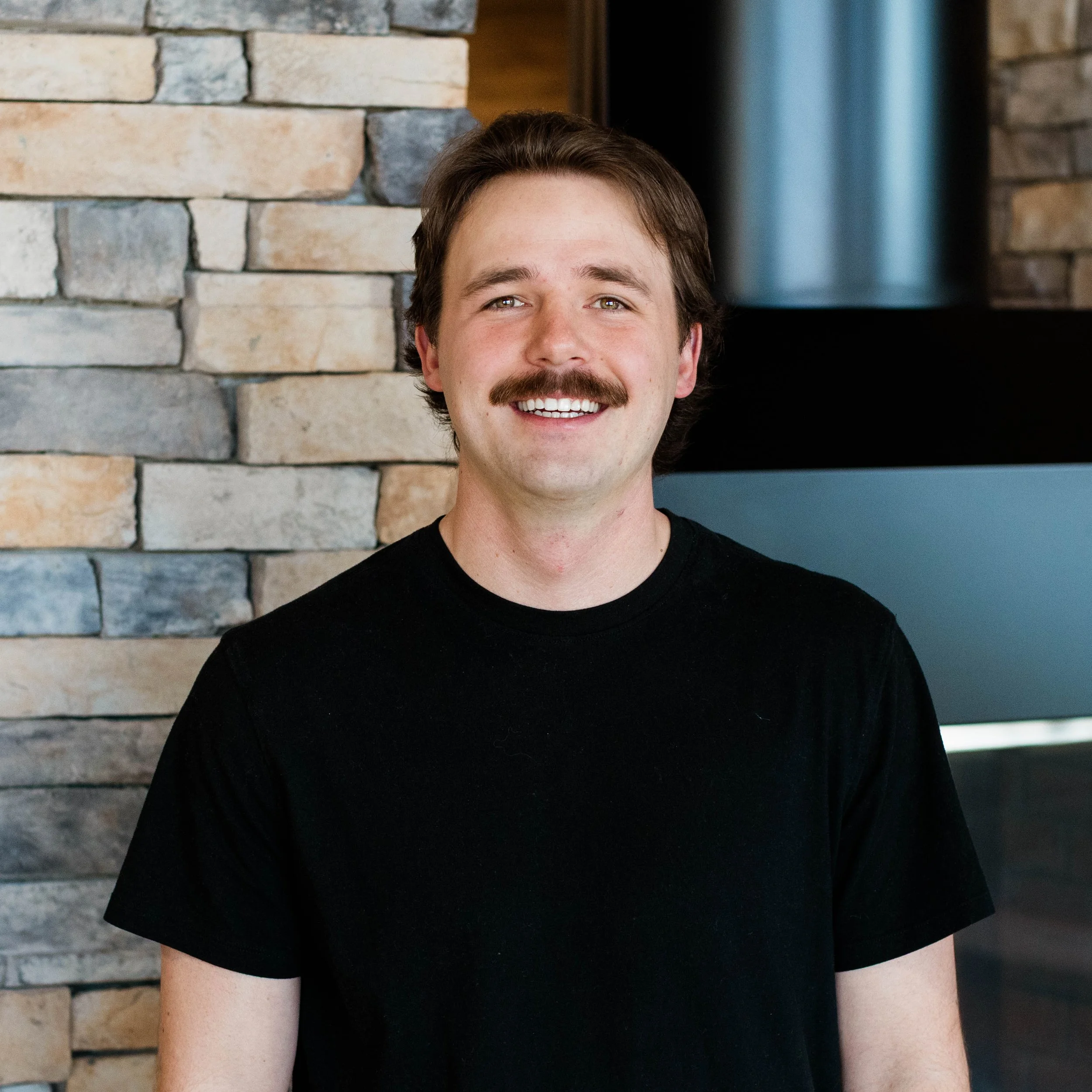 Smiling man with a mustache and dark hair, wearing a black t-shirt, standing in front of a stone wall near a fireplace.