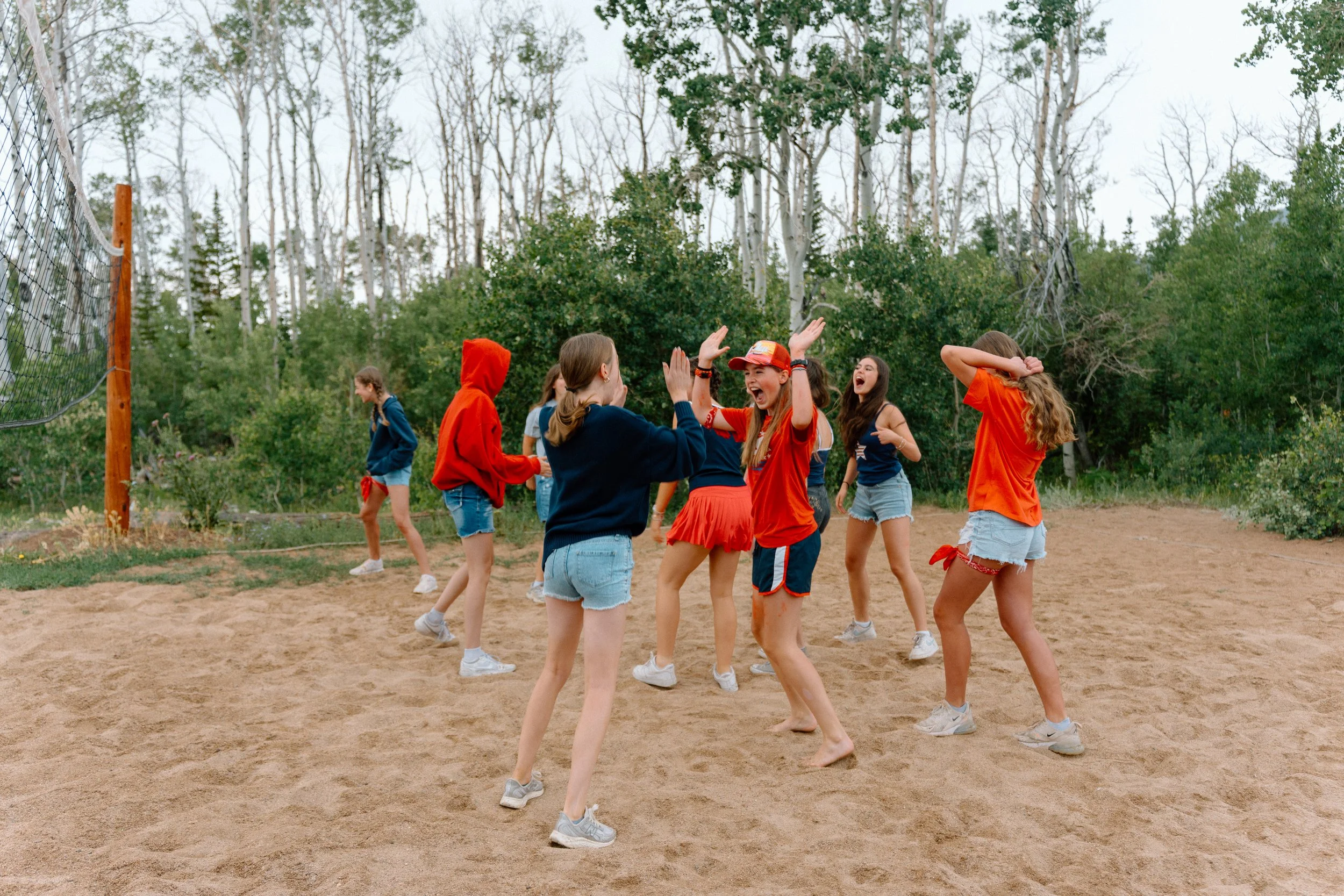Group of young girls playing volleyball outdoors on a sandy court surrounded by trees.