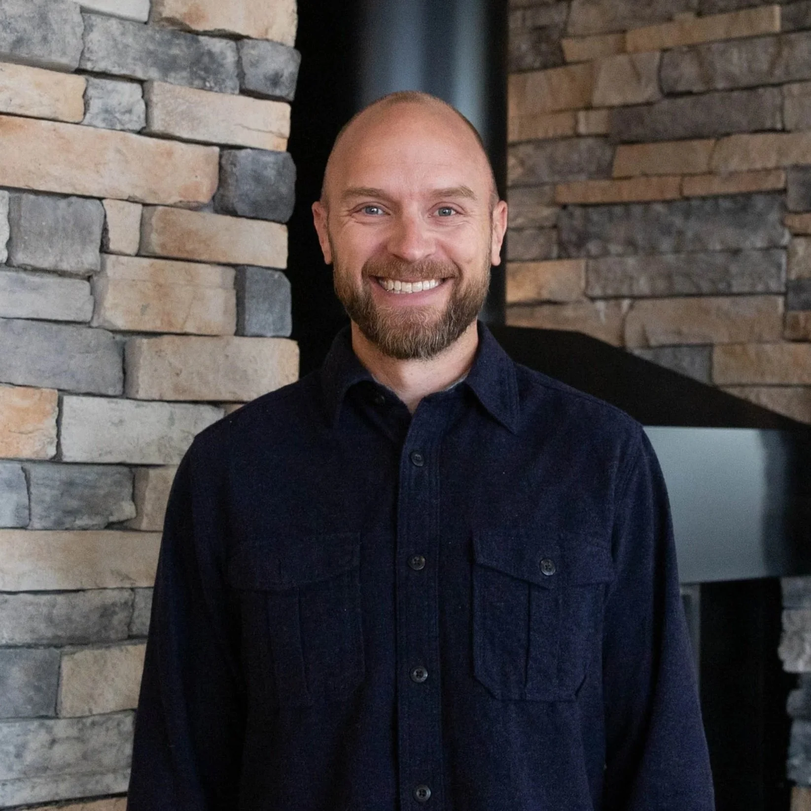 A smiling man with a beard and blue eyes standing in front of a stone wall.