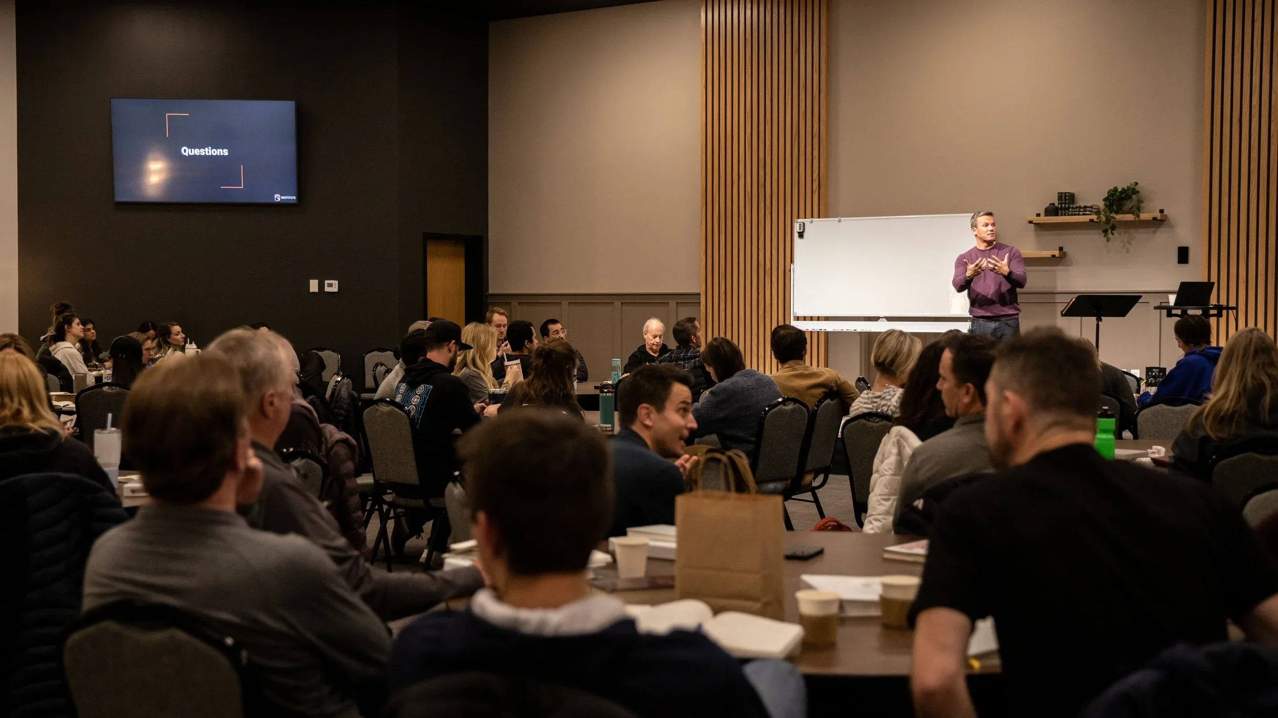 A man giving a presentation or lecture to a seated audience in a conference room, with a large whiteboard and a monitor displaying the word 'Questions'.