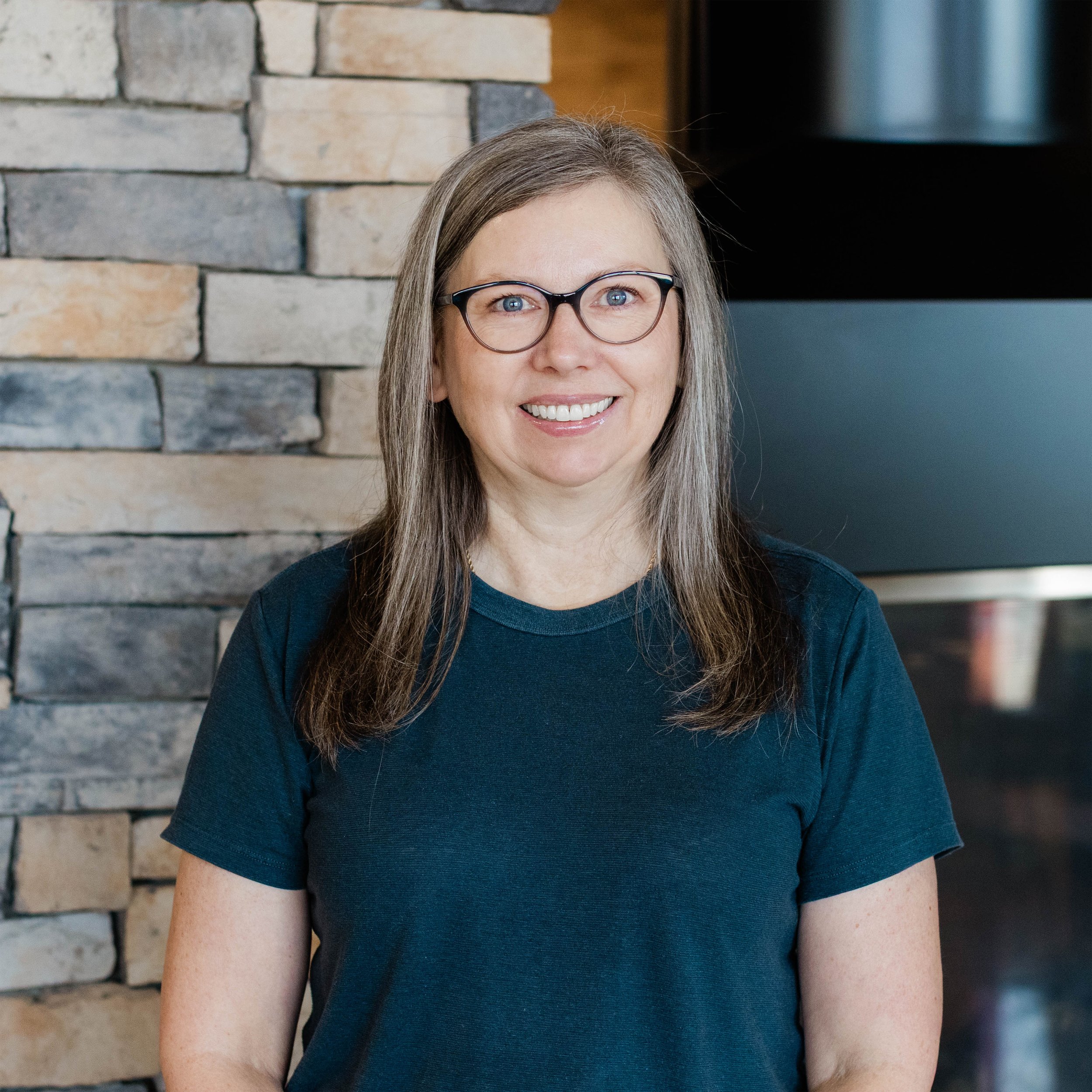 A woman with long brown hair, glasses, and a slight smile standing in front of a stone wall with a fireplace.