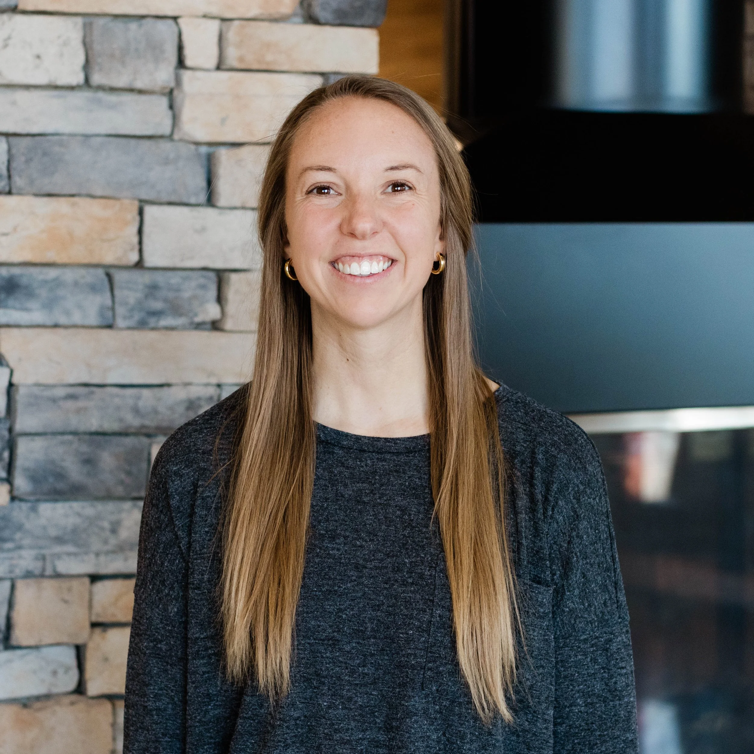 Smiling woman with long blonde hair standing in front of a stone fireplace wall.