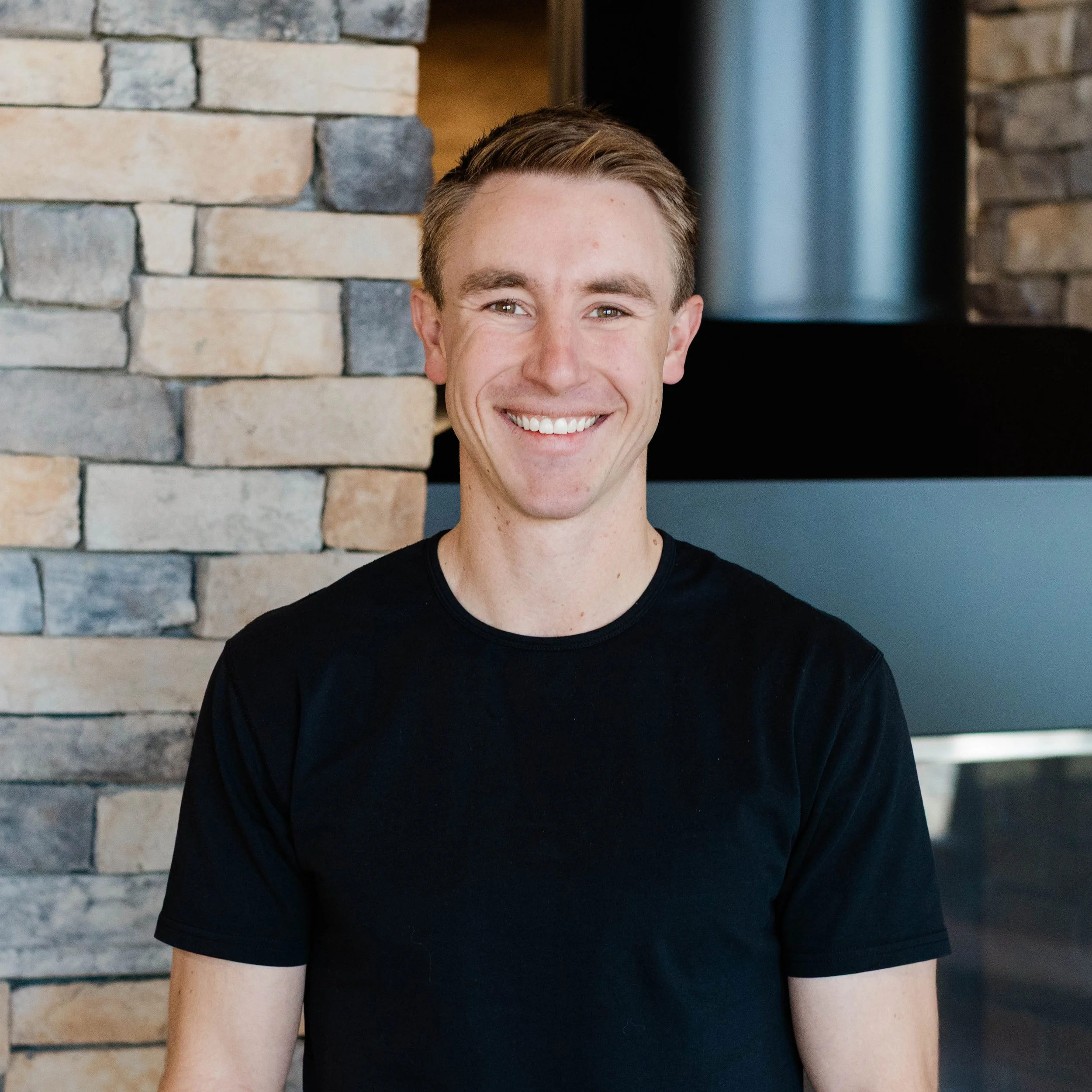 A young man with short brown hair and a big smile, wearing a beige polo shirt, standing in front of a stone wall with a fireplace.